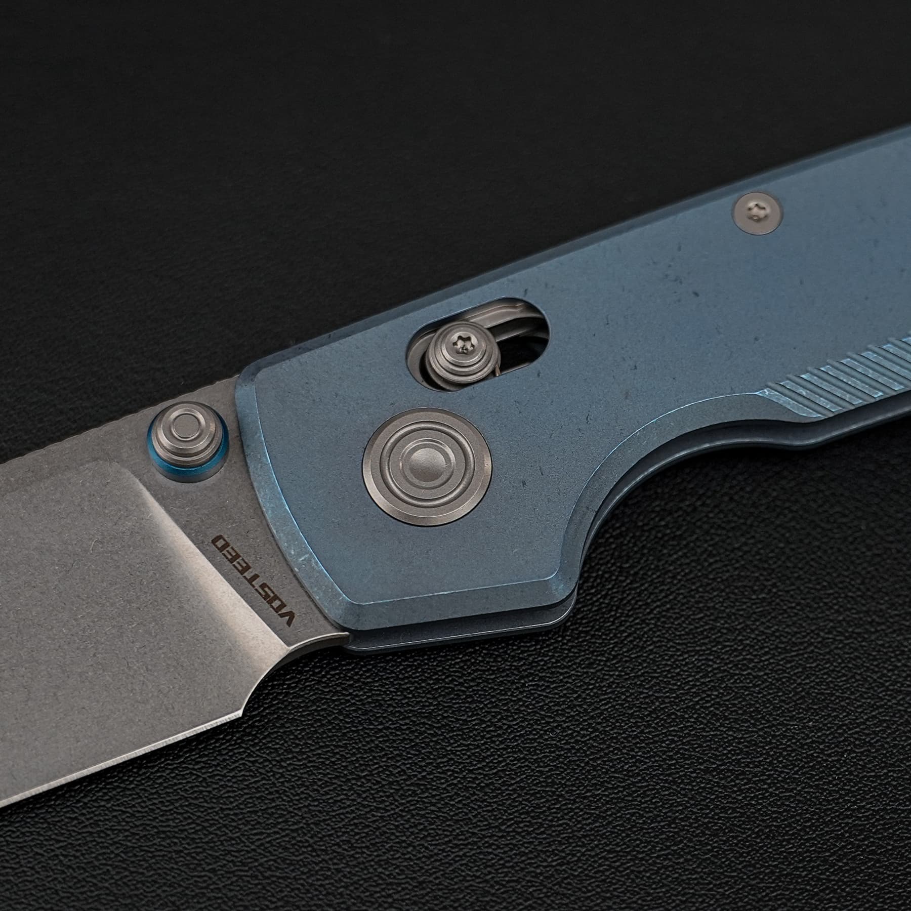 Close-up of a blue metal hinge with round rivets on a dark textured surface, likely part of a folding tool or multi-tool.