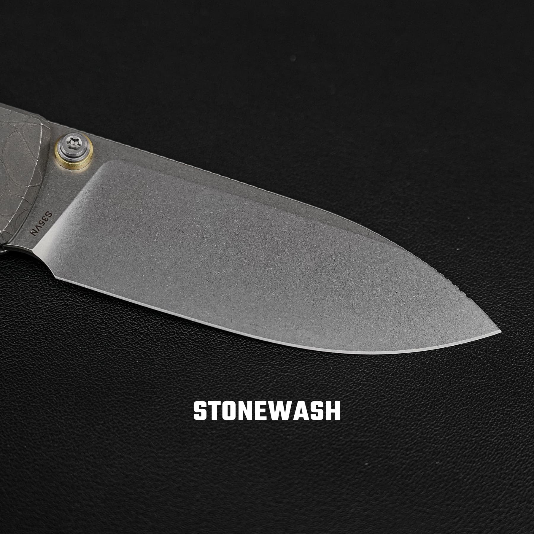 Close-up of a gray folding knife blade against a black textured background, with the word STONEWASH at the bottom.