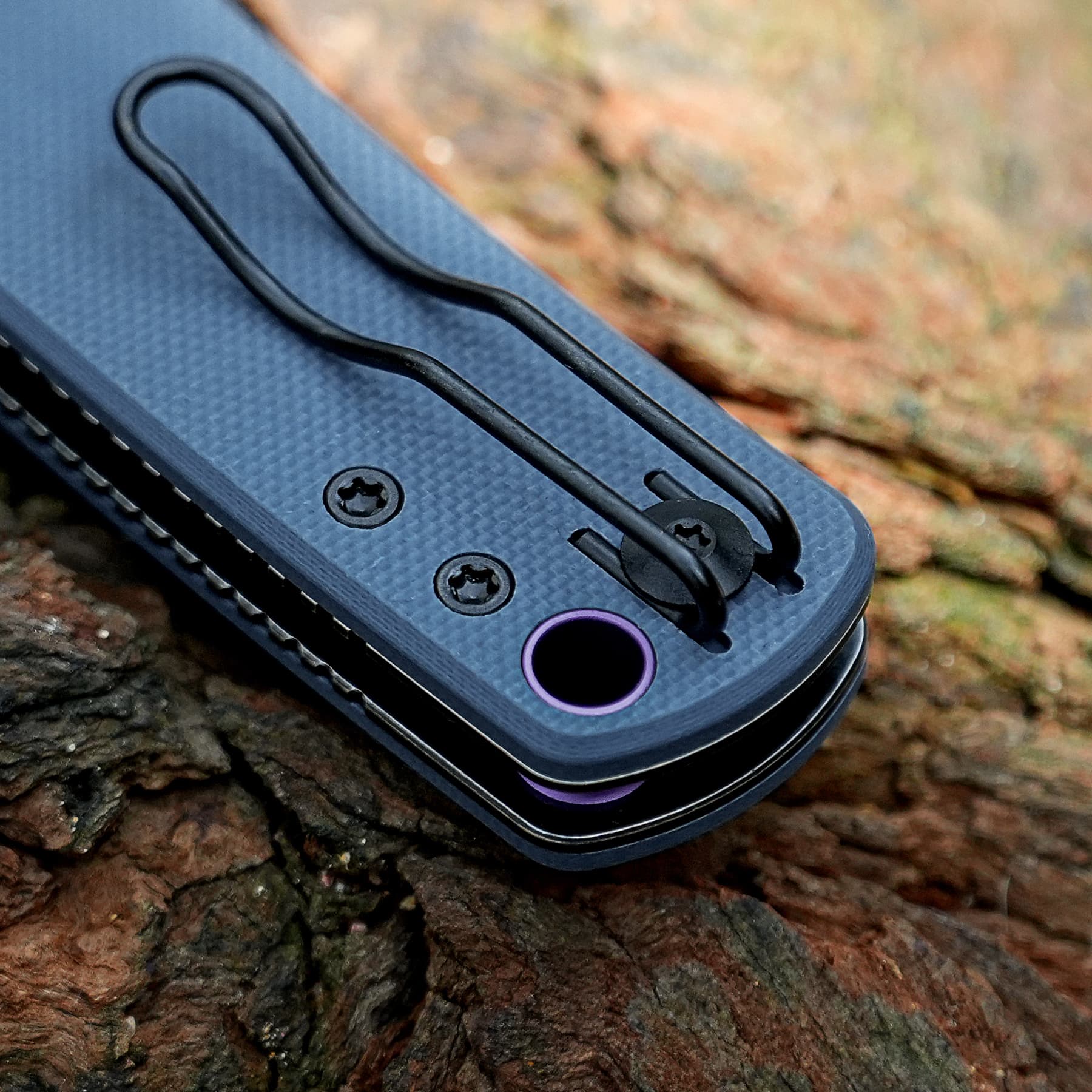 Close-up of a blue textured outdoor tool handle with screws and a black lanyard loop, resting on tree bark.