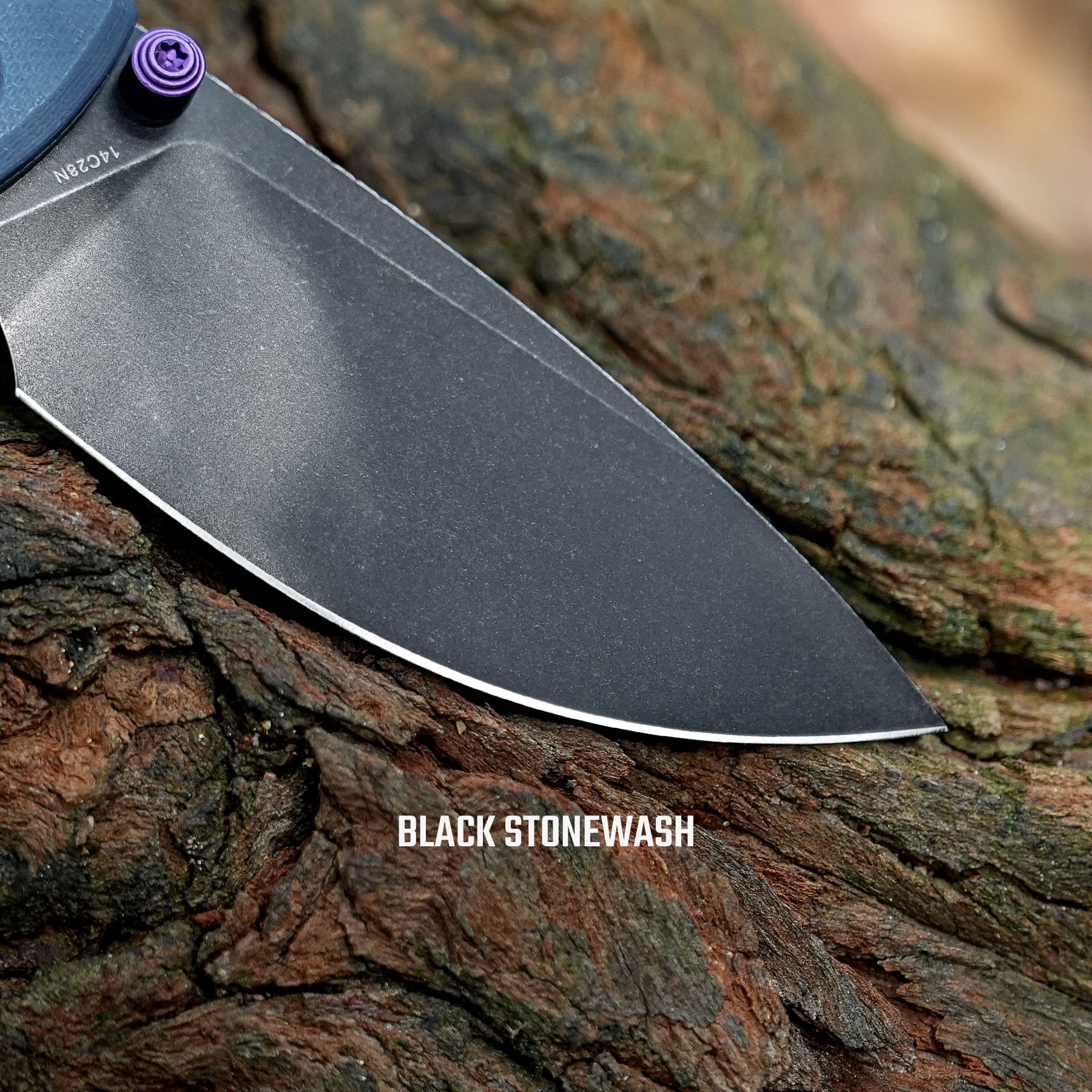 Close-up of a black stonewashed knife blade resting on rough tree bark, with the text 'BLACK STONEWASH' visible at the bottom.