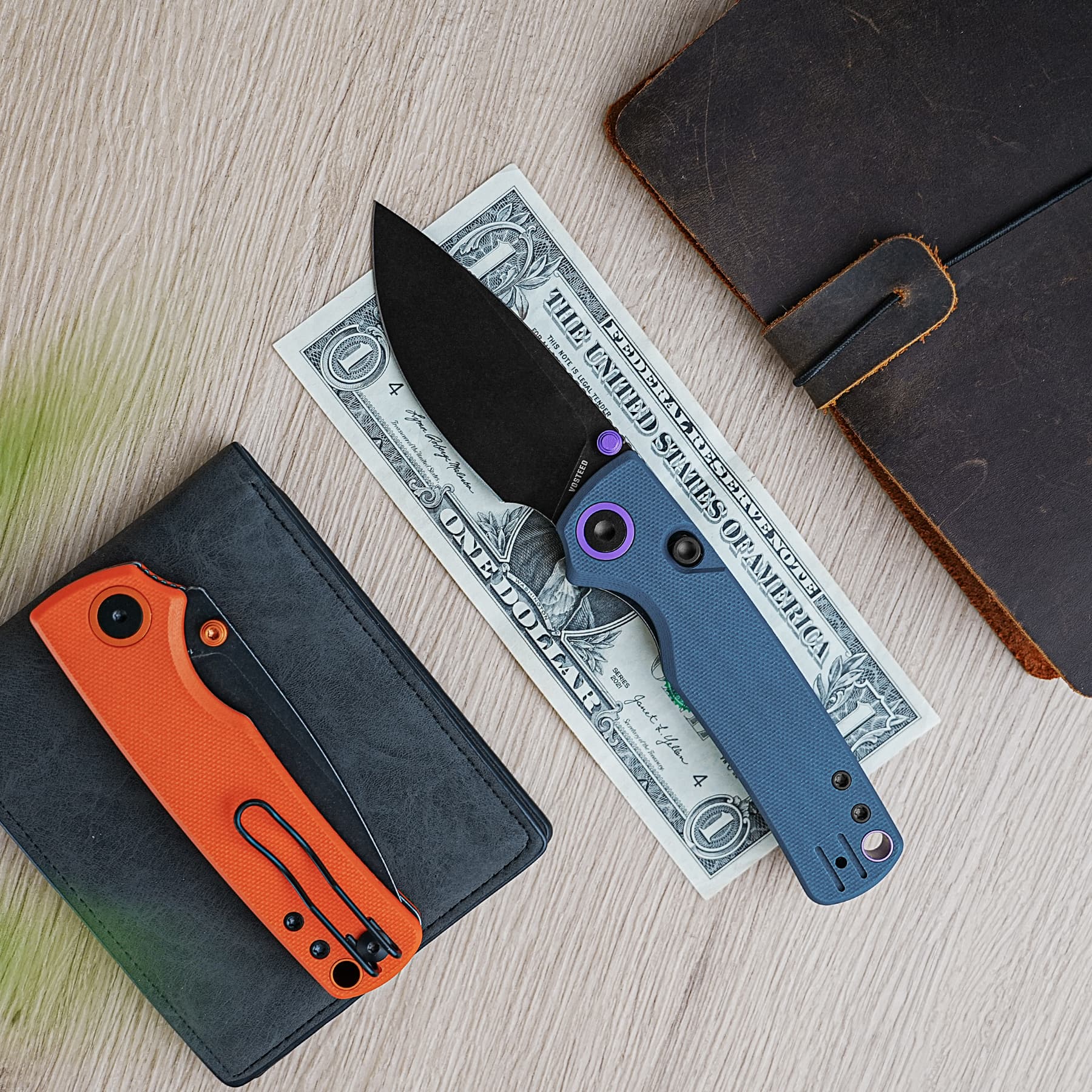 Close-up of two folding pocket knives tucked into black leather wallets on a wooden surface, with a one-dollar bill beneath the blades.