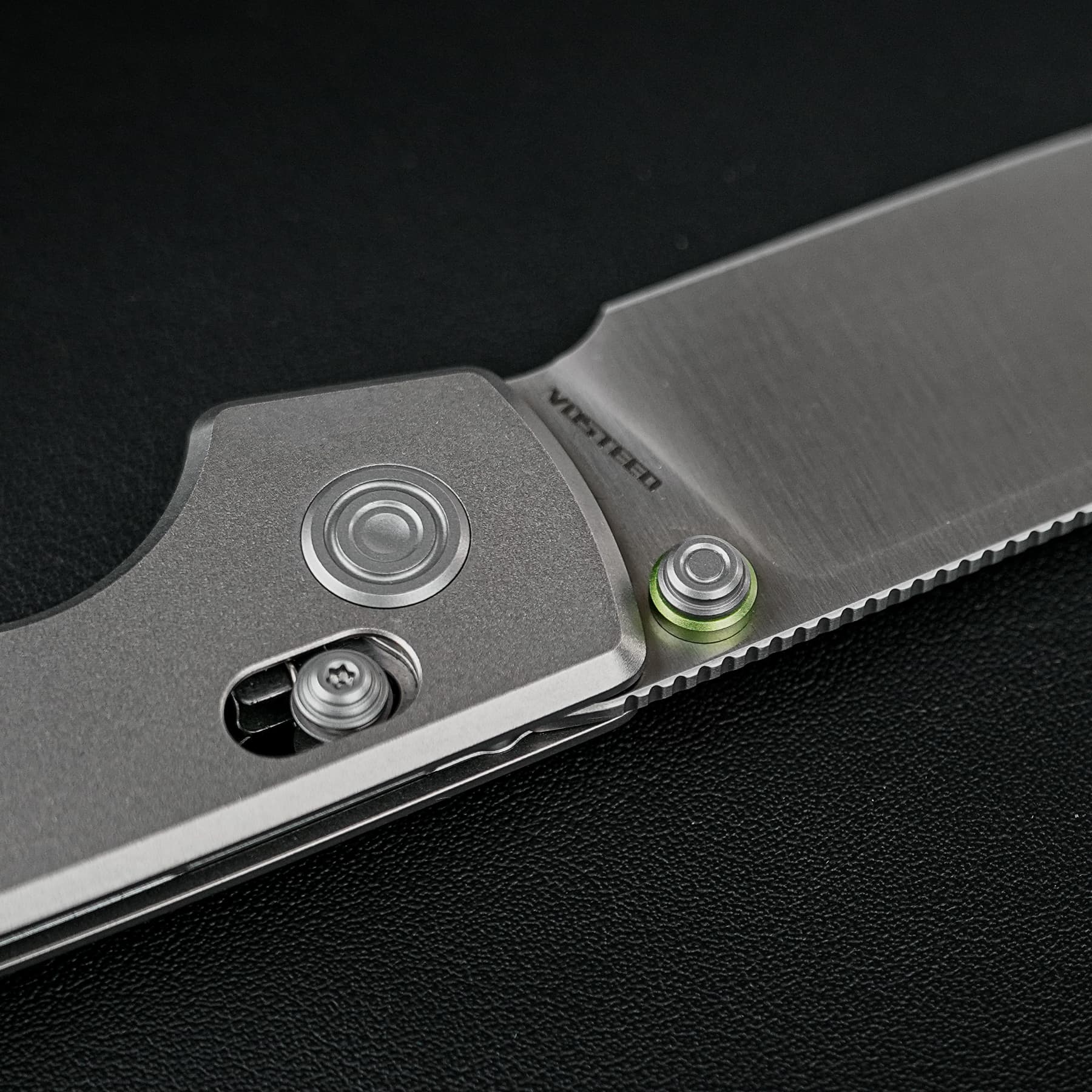 Close-up of a folding knife showing the brushed blade, exposed pivot and green accent ring on the handle against a dark background.