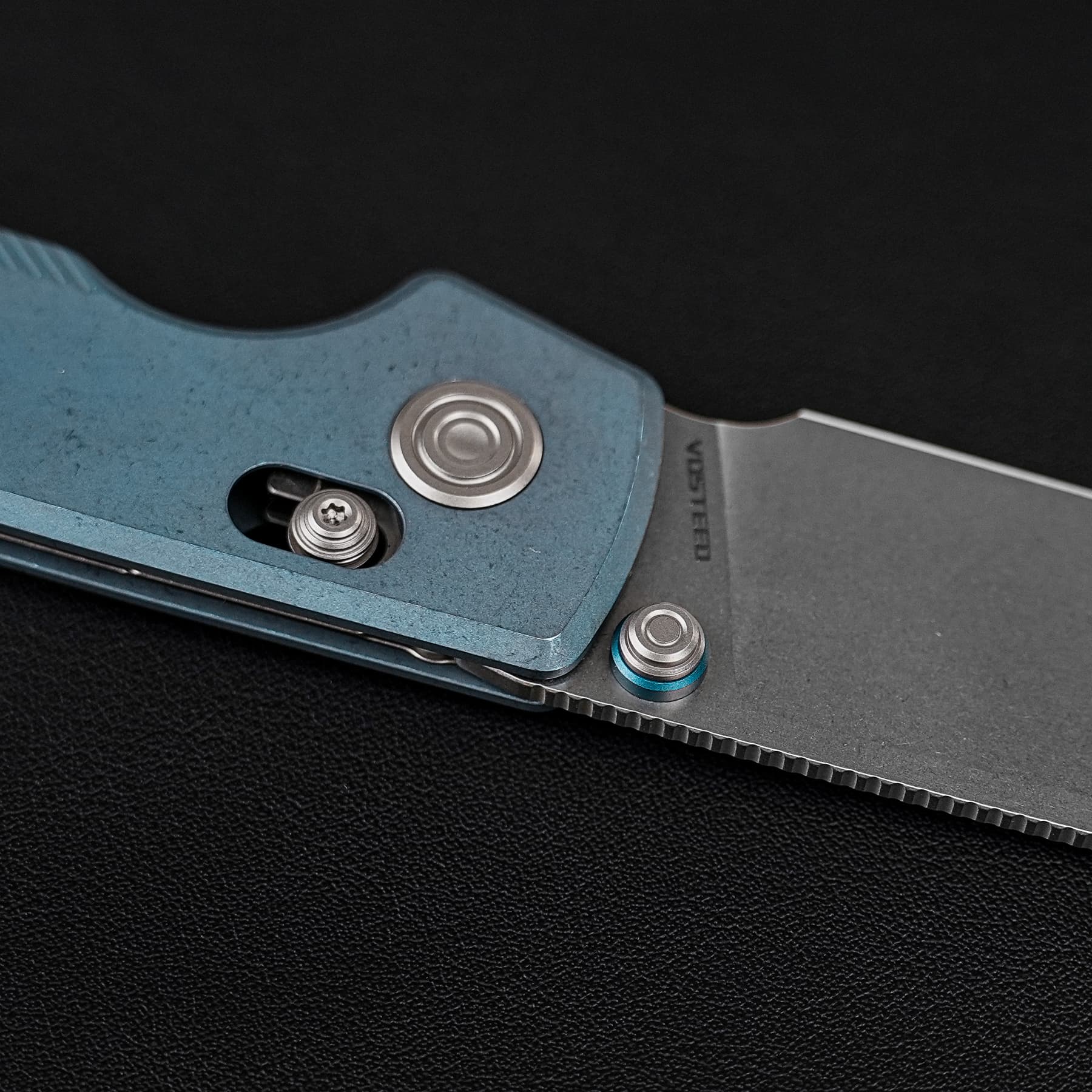 Close-up of a partially opened folding knife with a blue textured handle and metal blade on a black surface, showing the pivot and screws.