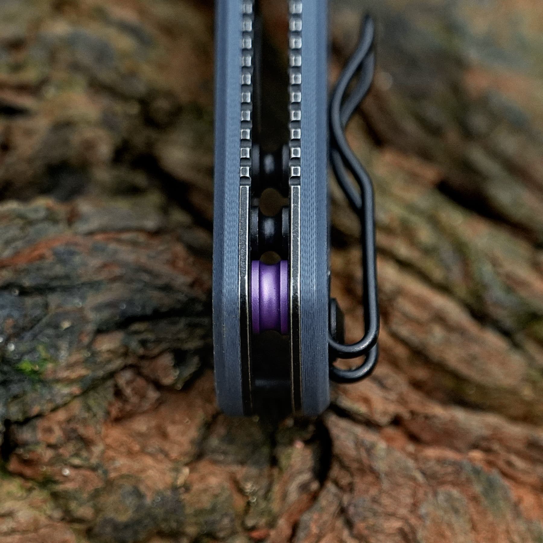 Close-up of a black device with two vertical rails and small square teeth along the sides, a purple circular component visible between the rails, and a looped cable on the right, set against rocky ground.