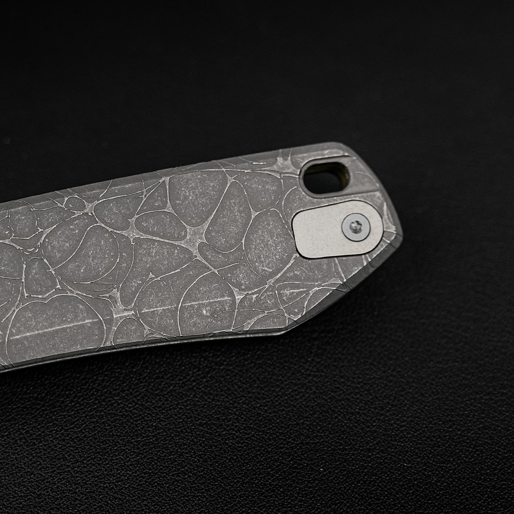Close-up of a patterned knife handle (stonewashed, marble-like) with a rectangular cutout and screw, set against a dark background.