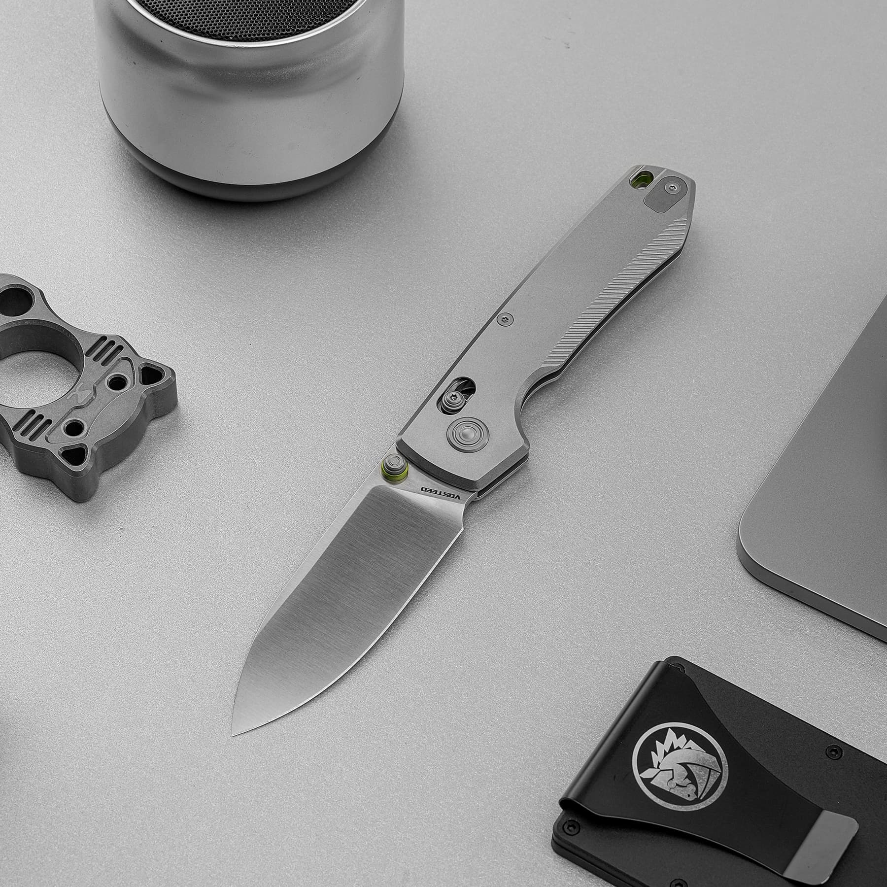 Folding knife with a textured handle and silver blade laid on a gray work surface, with metal components nearby.