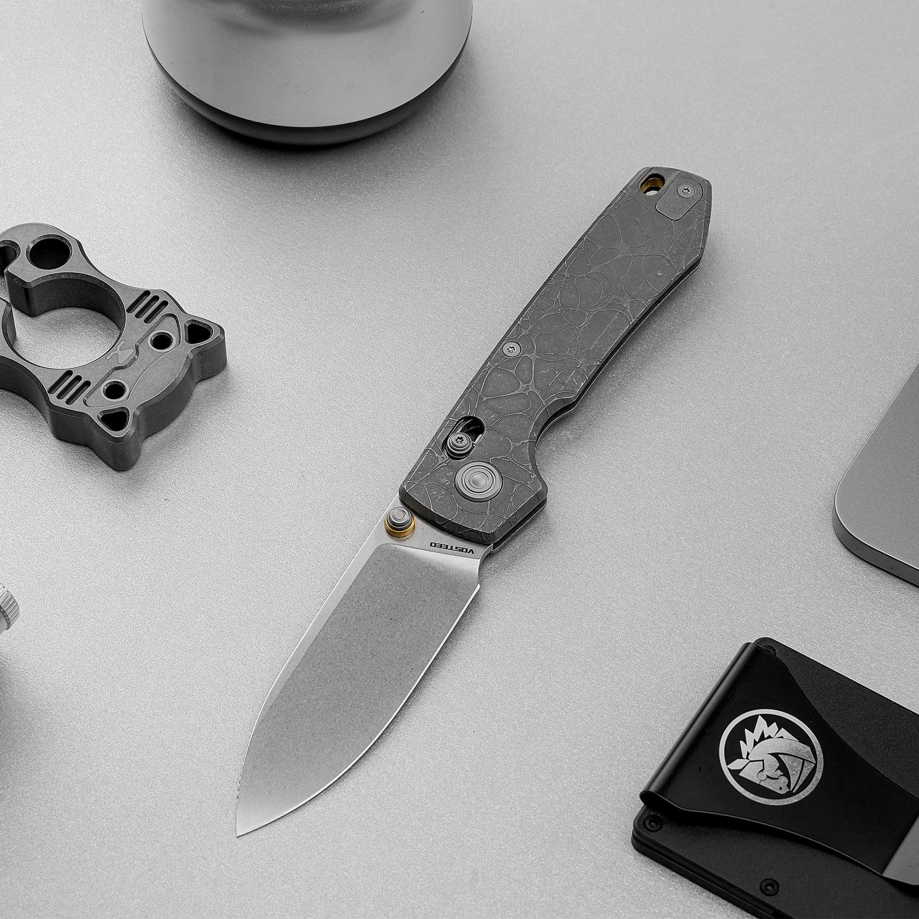 Close-up of a folding knife with a cracked-pattern handle and blade extended on a gray surface, beside a black logo-printed case.