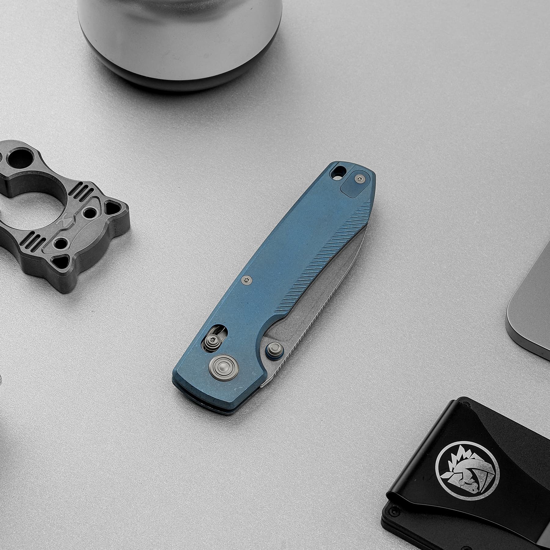 Blue retractable utility knife lying diagonally on a textured gray work surface near a metal clamp component.