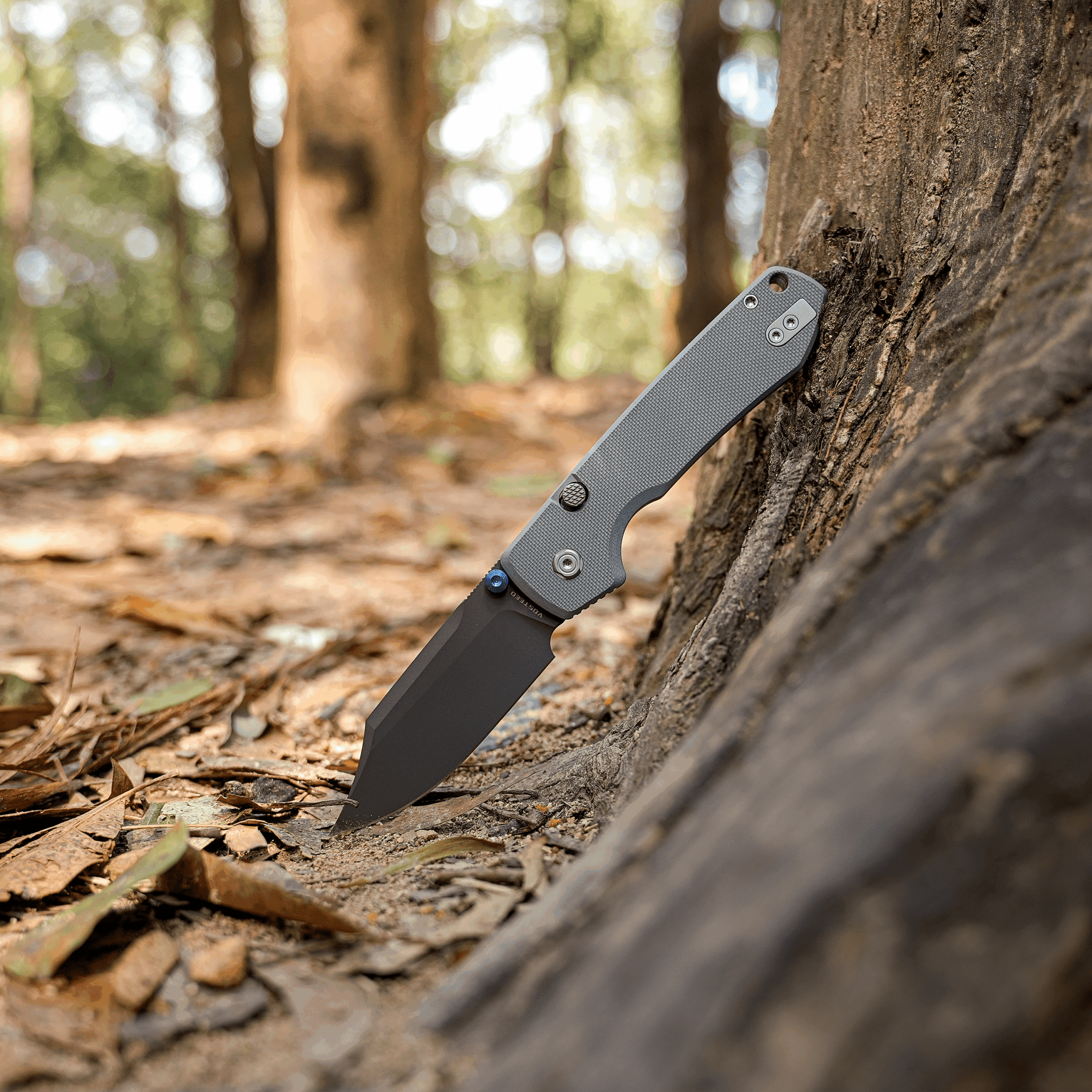 Gray folding knife stuck into a tree trunk with a blurred forest background behind it, outdoors.