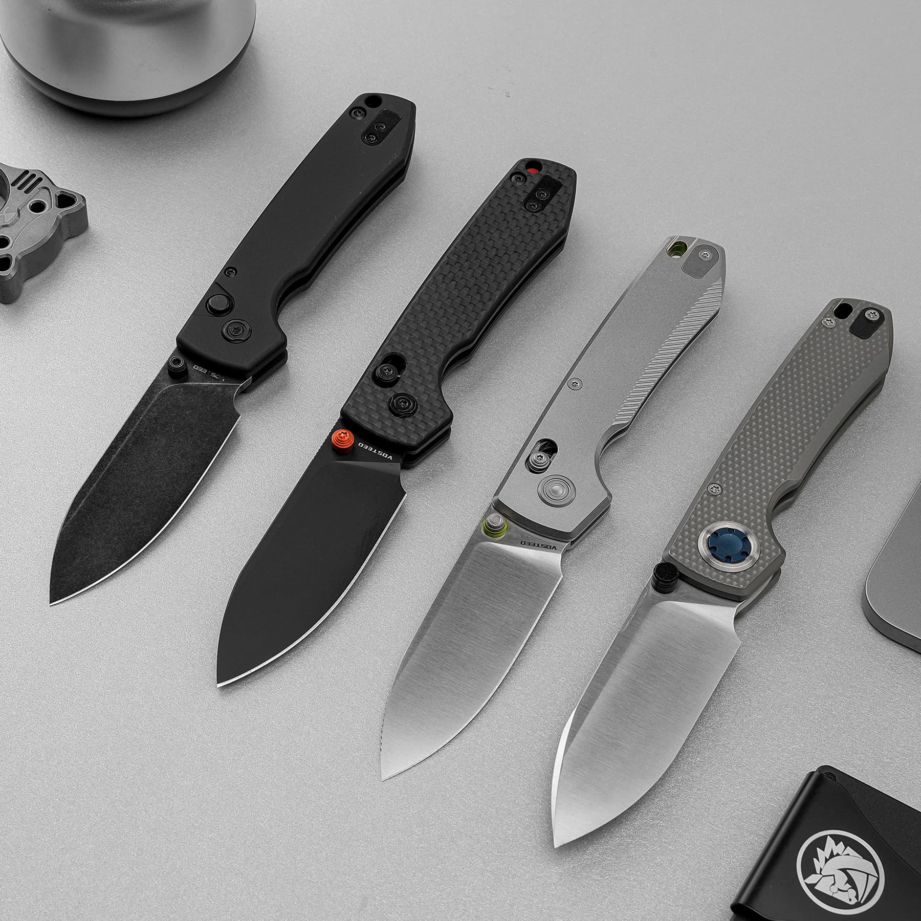 Five folding knives with metal blades and textured handles laid diagonally on a gray surface, showing varied designs and finishes.