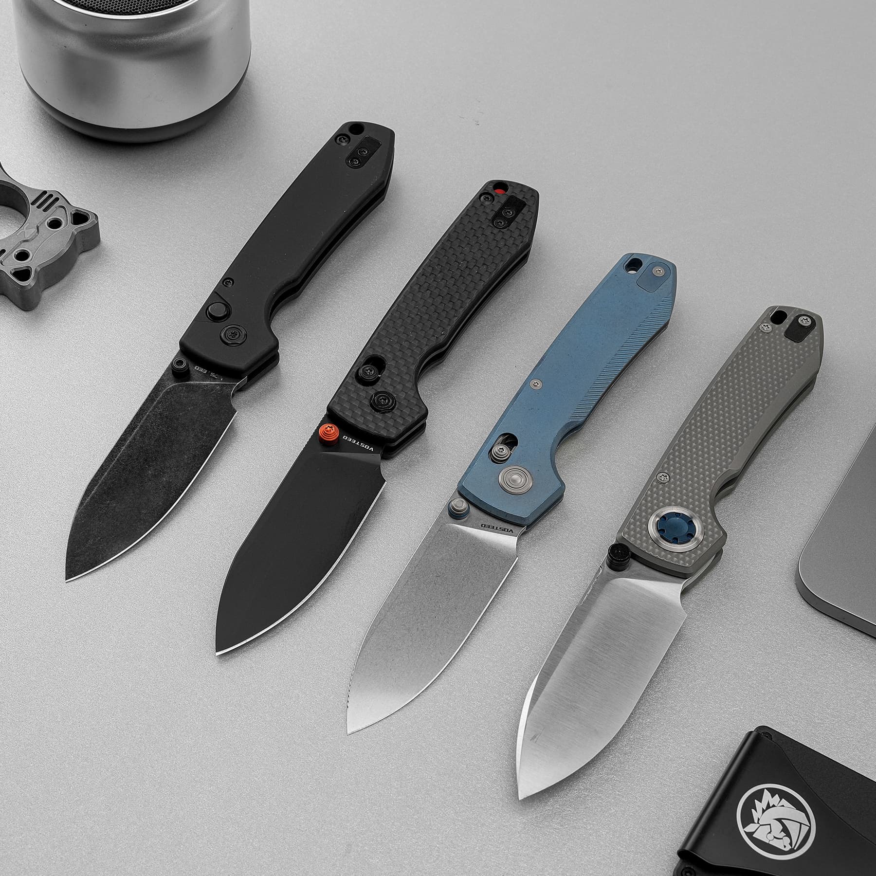 Row of folding knives with varied handle colors (black, carbon fiber, blue, gray) and exposed blades on a gray surface, showcasing their hardware and design.
