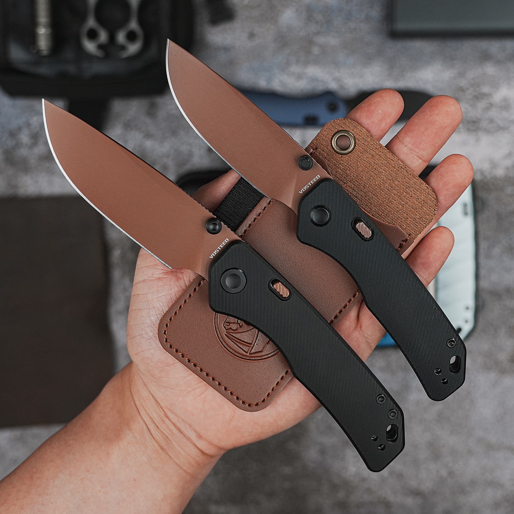 Two tan and black folding knives with brown textured handles held in a hand above a gray surface, with leather sheath visible nearby.