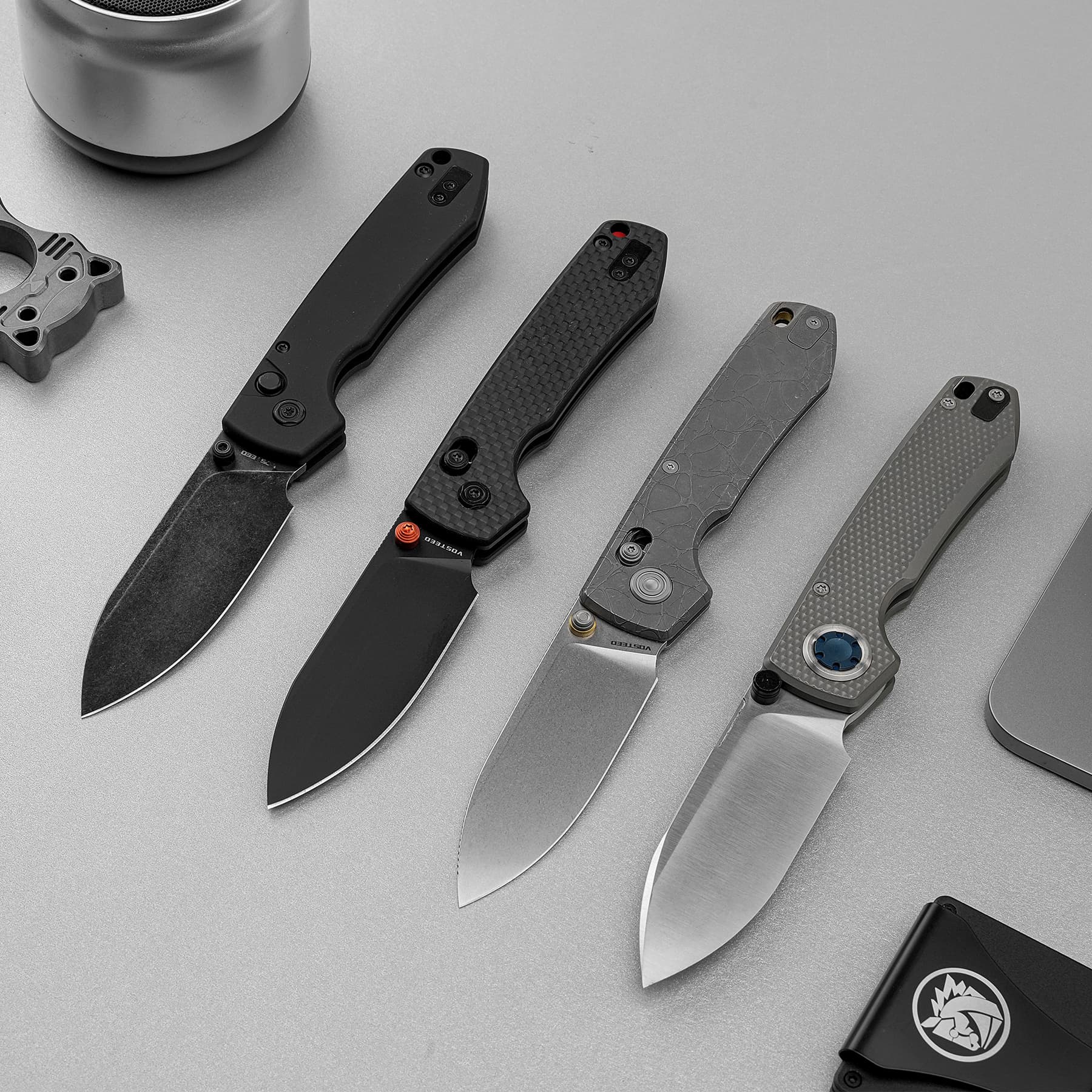 Collection of four folding knives with textured handles laid out diagonally on a gray surface, showing different blade finishes and hardware details.