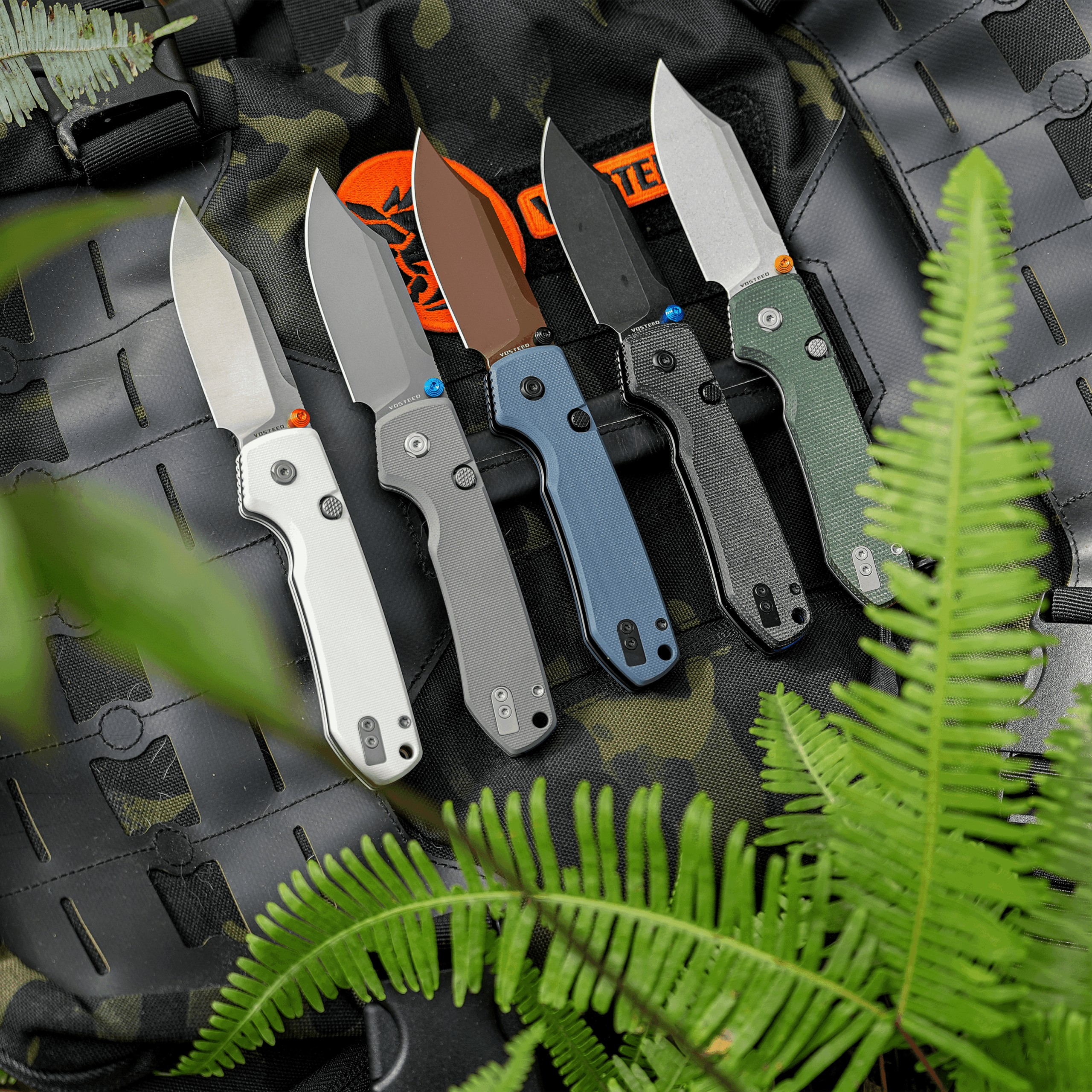 Knives with colored handles mounted on a camouflage tactical vest, framed by green fern fronds in a outdoor gear setup