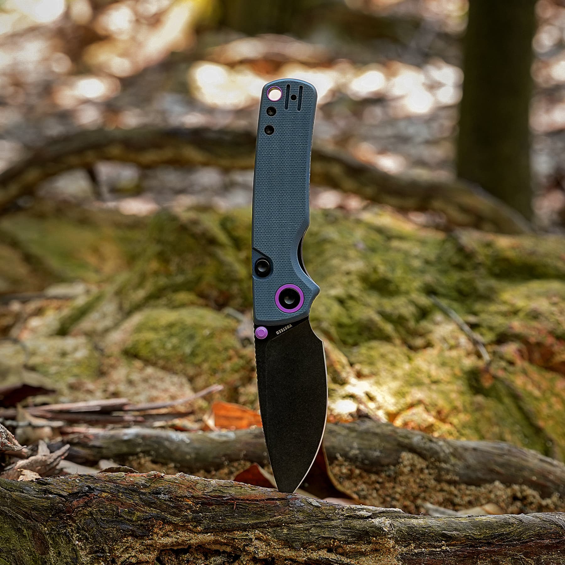 Close-up of a gray folding knife with purple hardware, blade partially open, embedded in a mossy log in a forest setting.