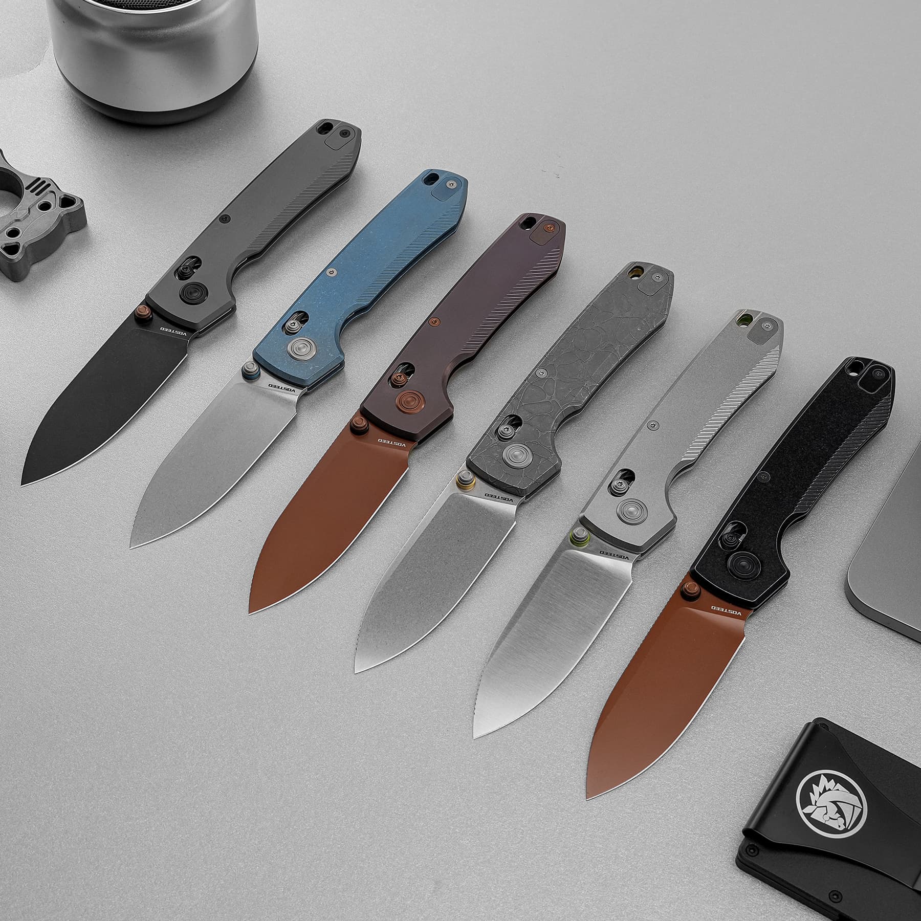 Row of folding knives with colored handles (gray, blue, brown, marbled, black) on a gray surface, showing different blade styles and hardware, top-left partially visible bottle cap accessory.