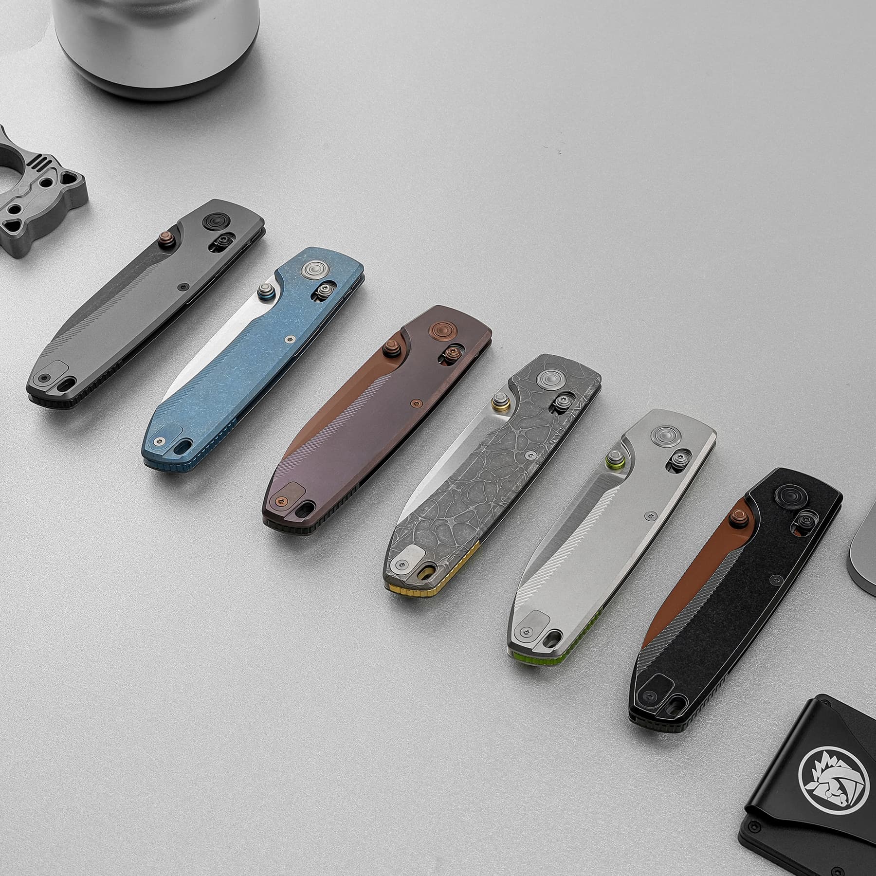 Several folding knives with various colored handles (blue, brown, gray, black) lie diagonally on a light gray surface, blades closed and visible hardware evident on each.