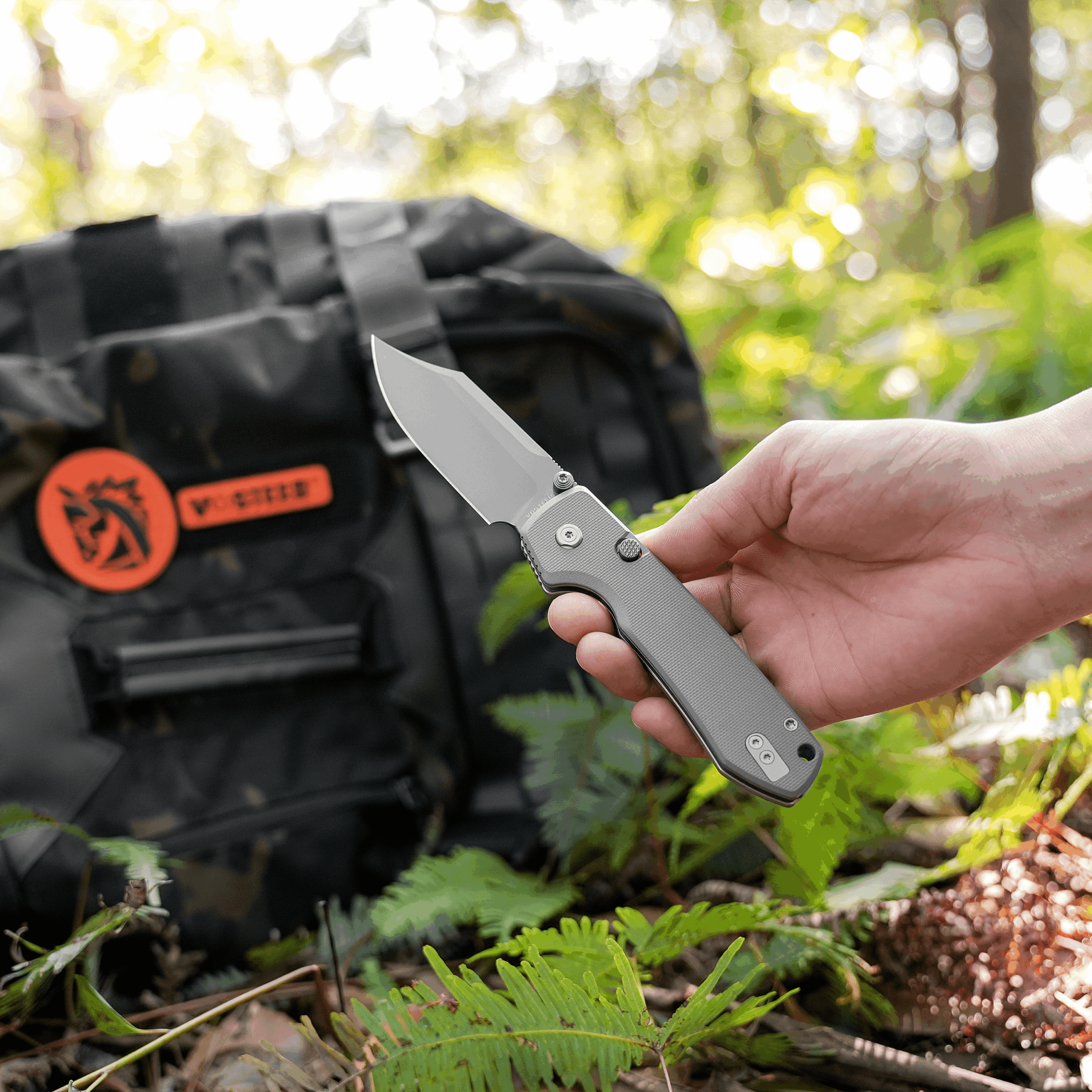 Close-up of a hand holding a folding knife outdoors, with a blurred backpack and green foliage in the background.