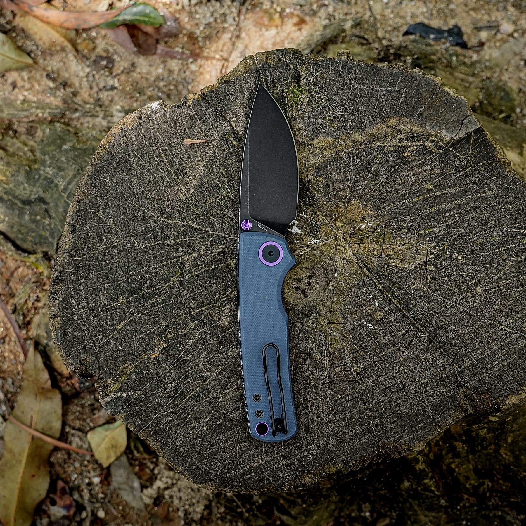 Blue folding knife with a black blade resting on a weathered tree stump outdoors.