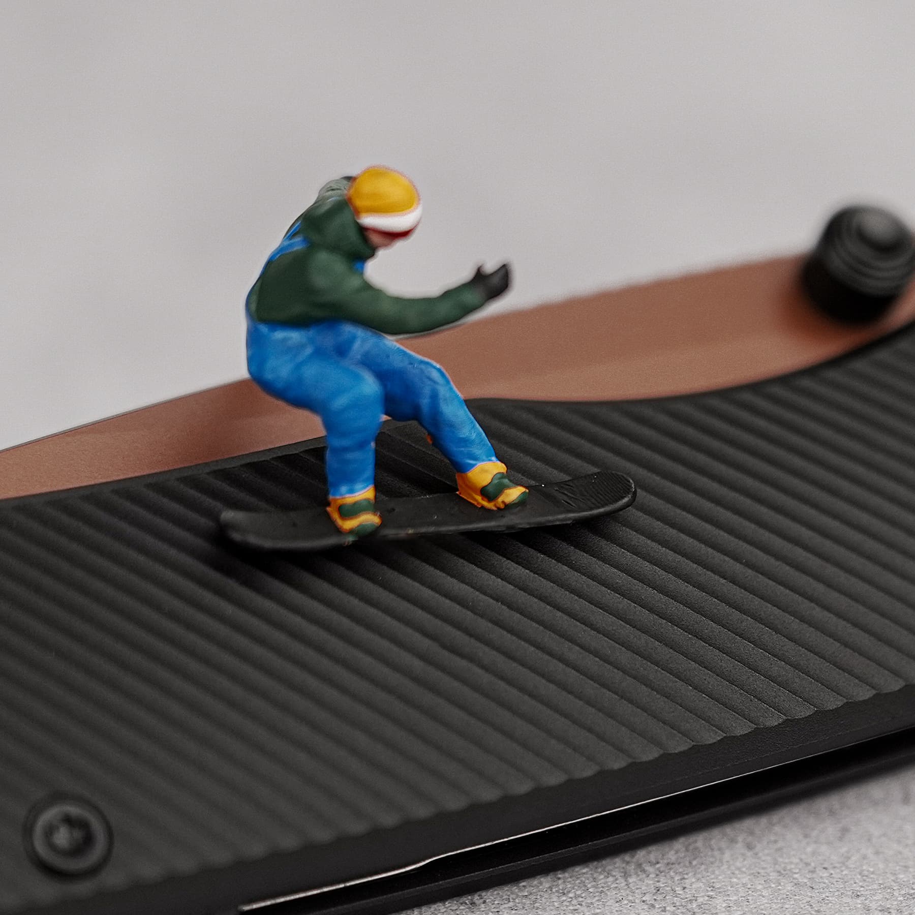 Small toy figure wearing a green top, blue pants, and a yellow helmet, posed on a black ribbed surface as if skating or surfing a slope of texture.