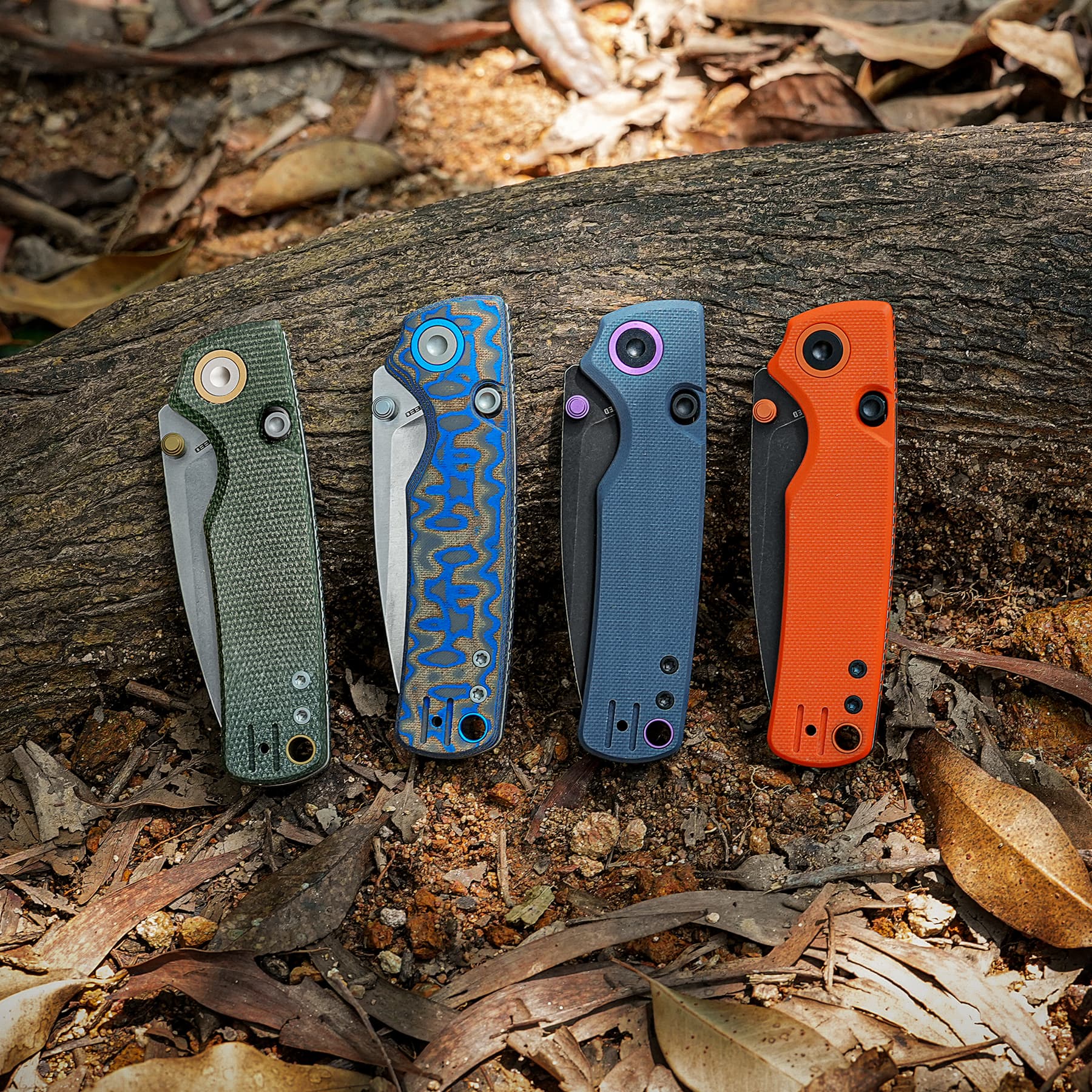 Five colorful folding knives with patterned handles lined up on a rough wooden log outdoors.