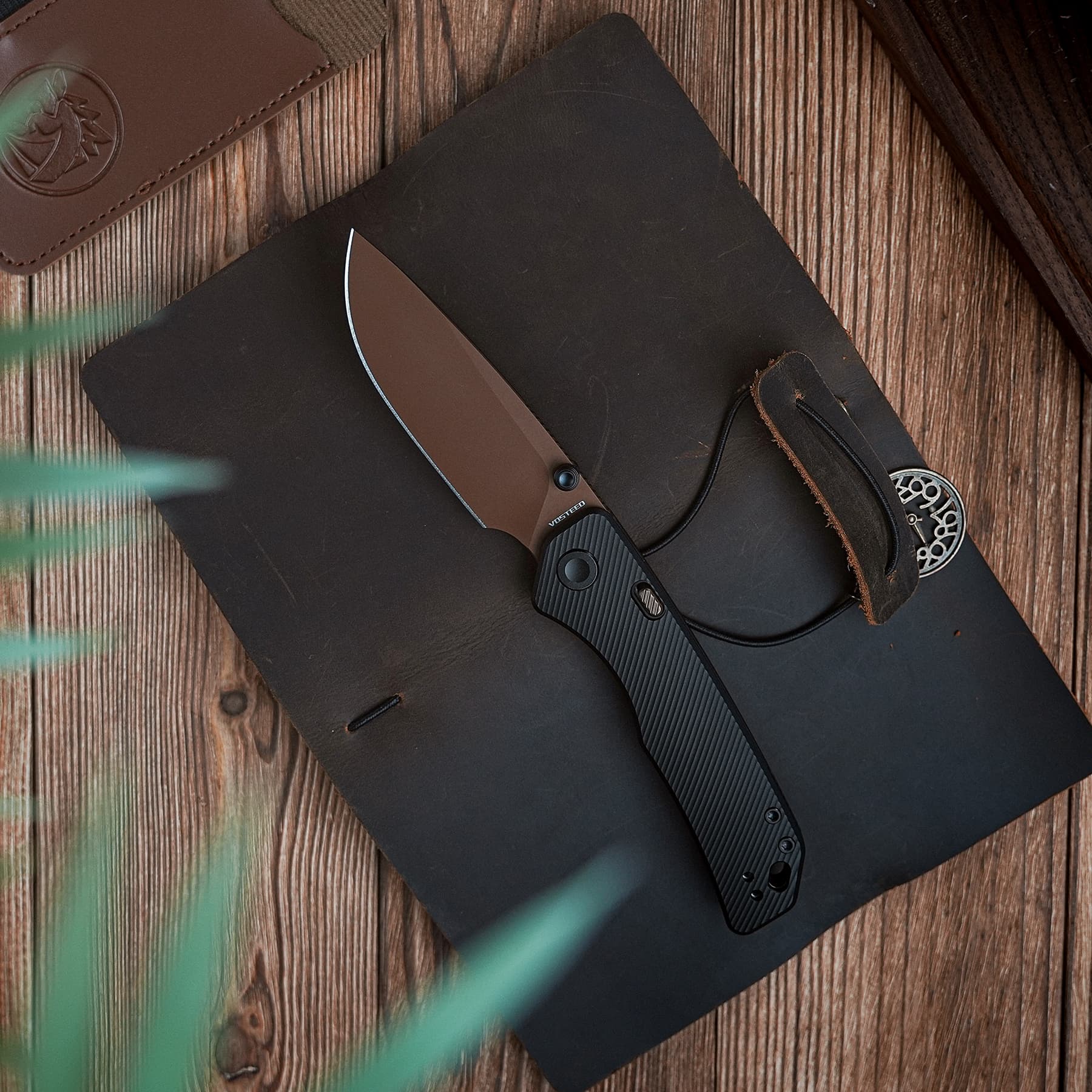 Folded black-handled knife with brown blade resting on a dark notebook on a wooden table at an angle.