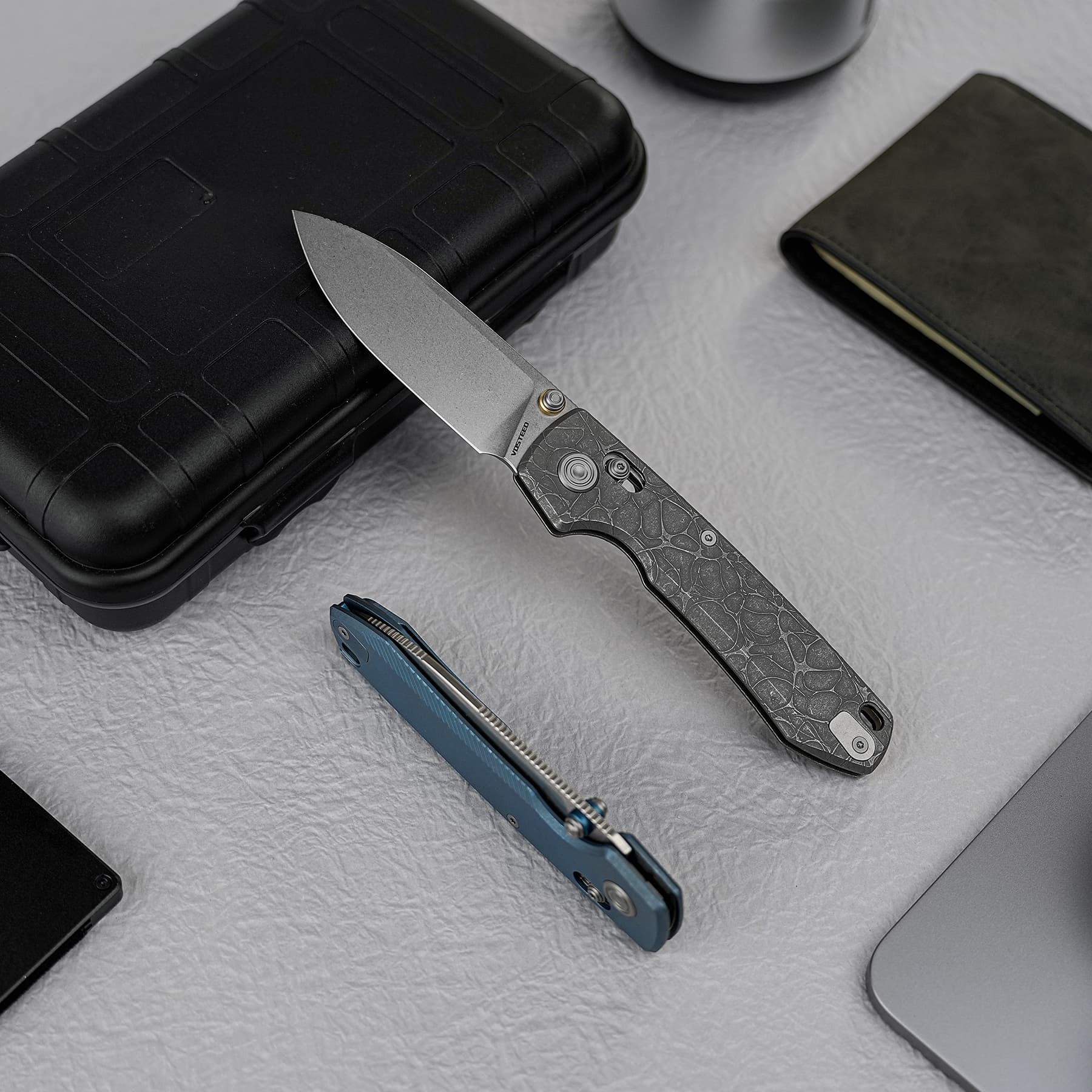 A partially opened folding knife with a textured handle placed on a black protective case on a gray surface.