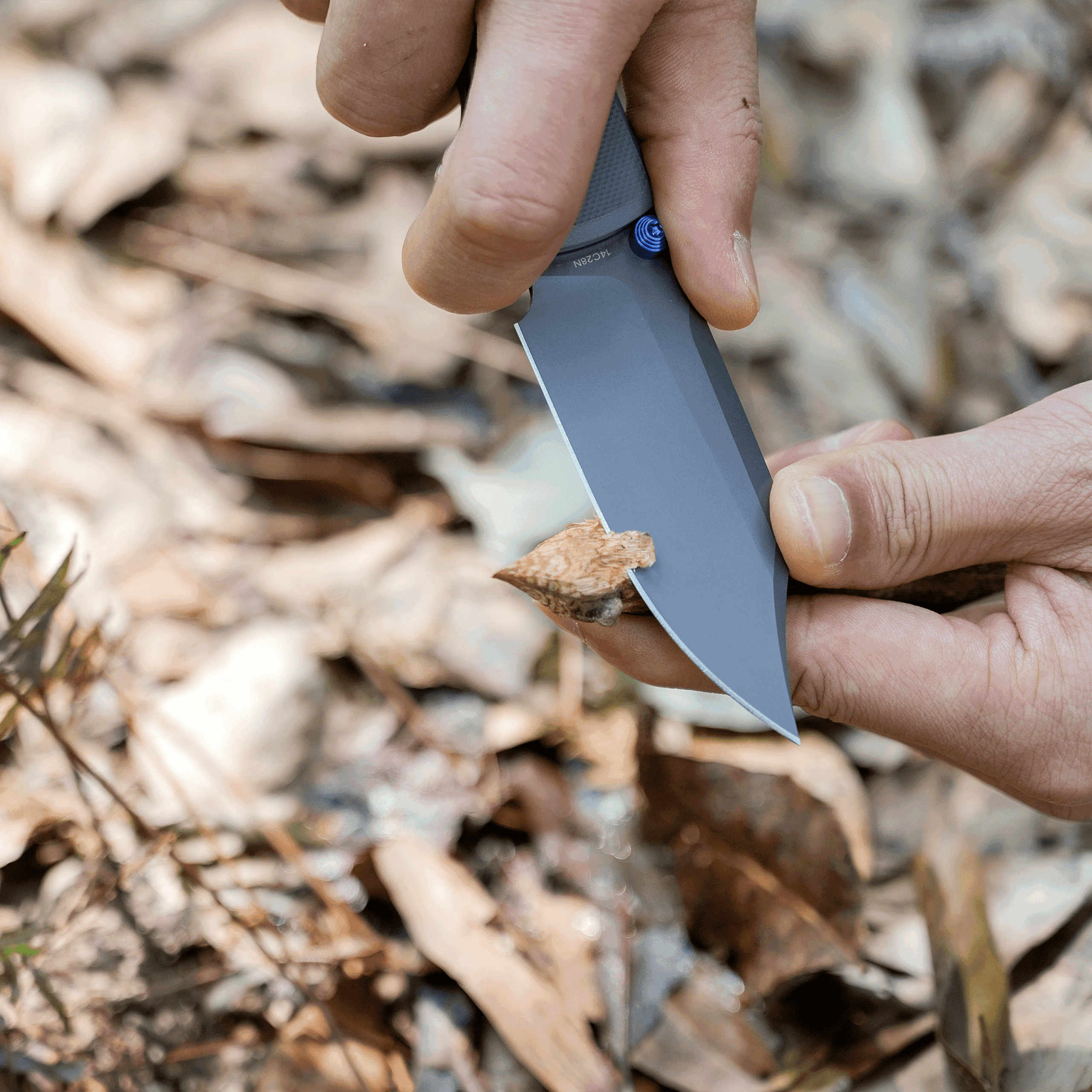 Close-up of a person’s hands holding a gray blade, sharpening it against a tool outdoors with wood shavings nearby.