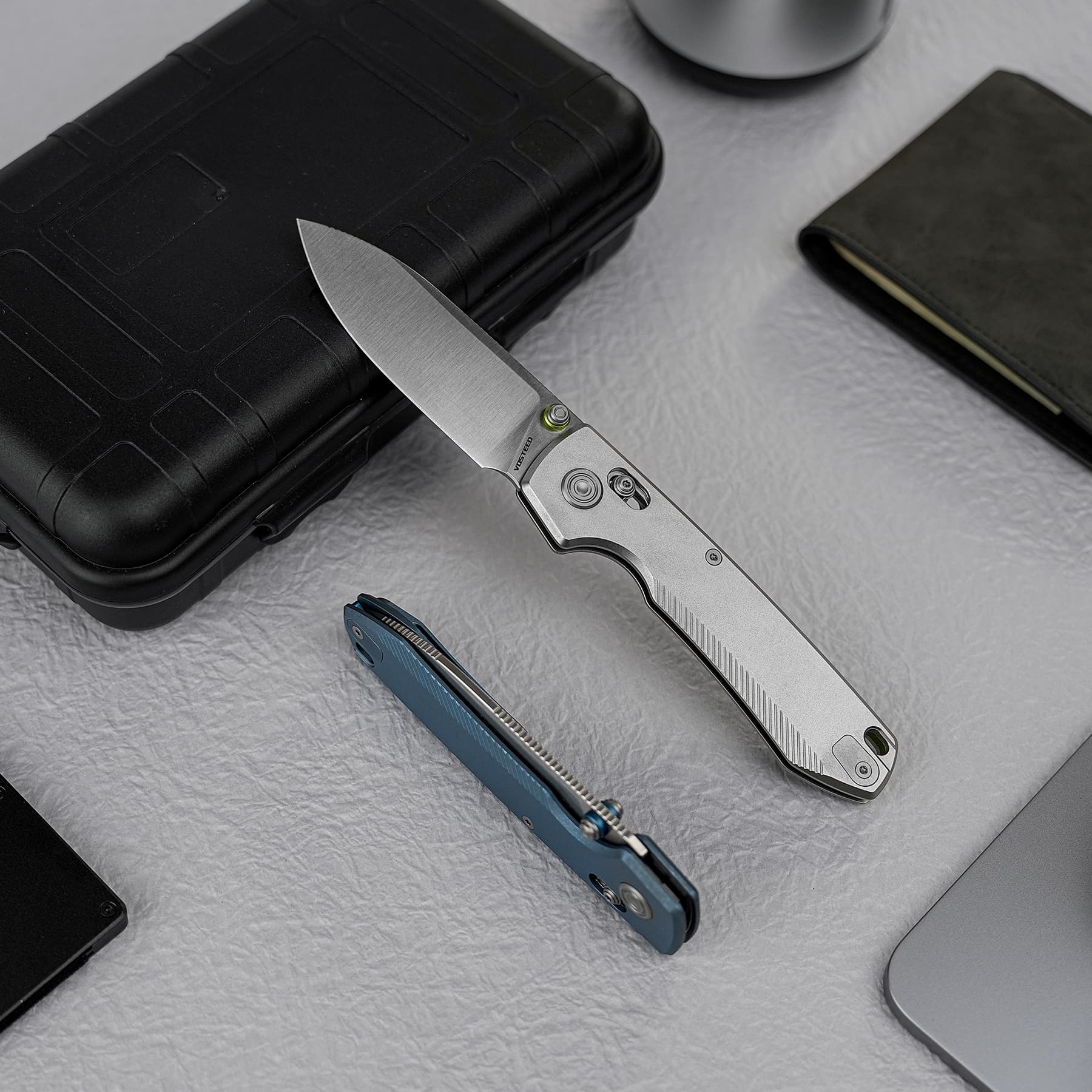 Open folding knife with a silver blade resting on a black protective case, with a blue multitool nearby on a gray surface.