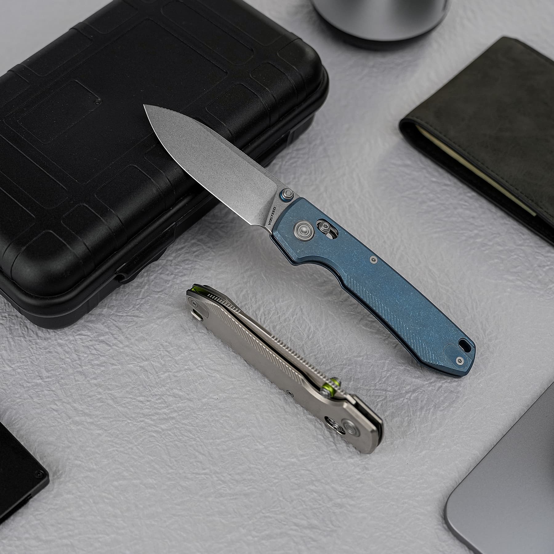 Folding knife with a blue handle partially opened on a textured gray surface beside a black protective case and a metal tool.