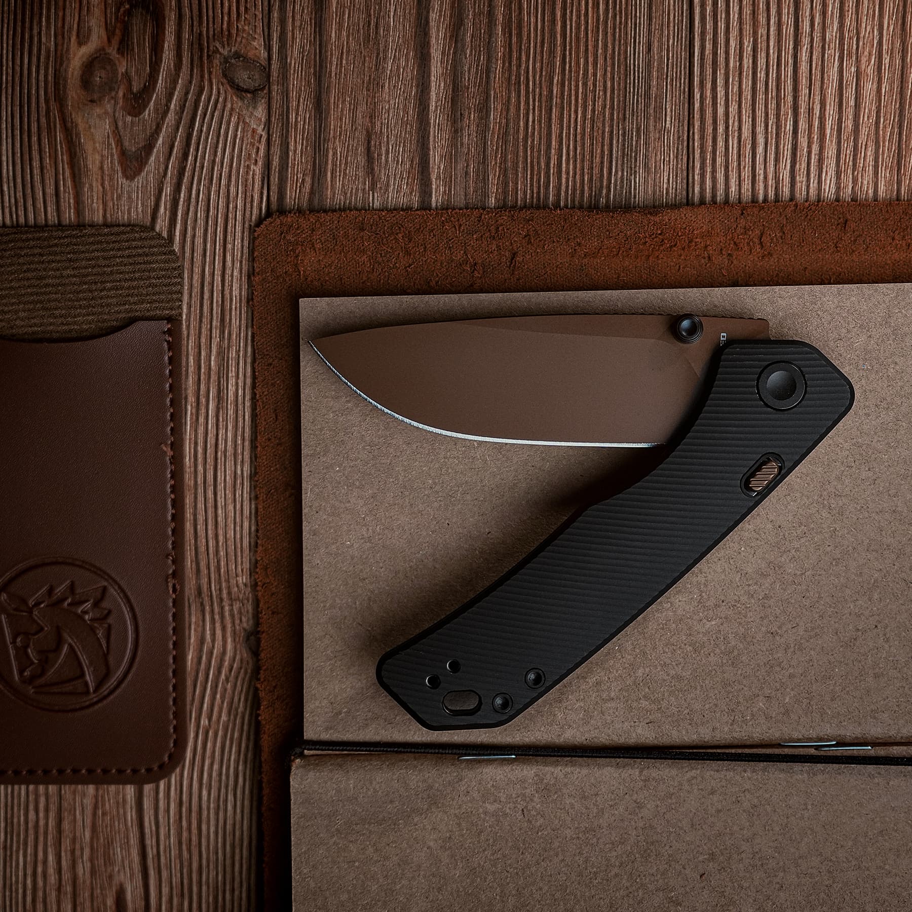 Close-up of a folding knife with a curved blade and black textured handle resting on a brown desk mat over a wooden surface.