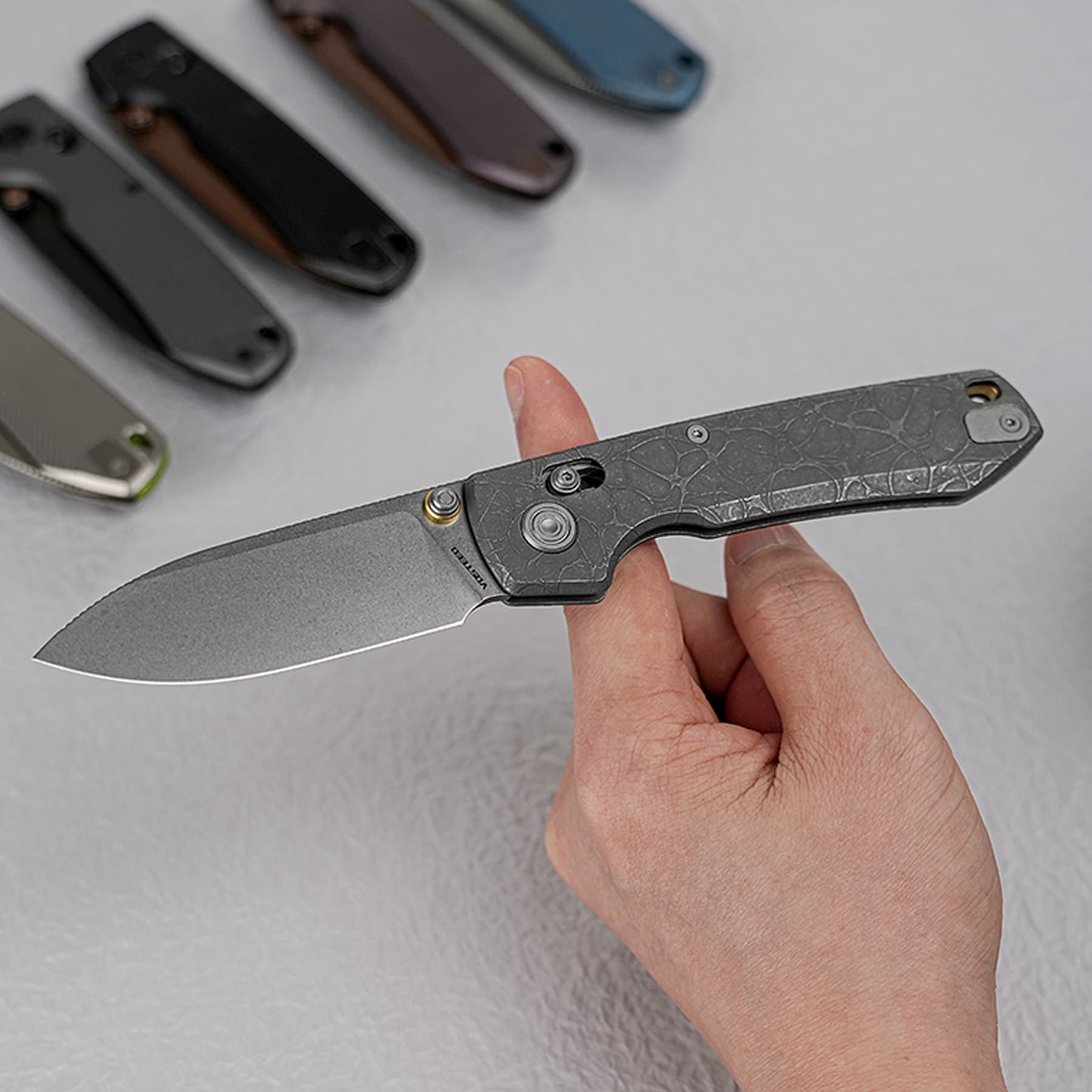 Close-up of a hand holding a folding knife with a gray patterned handle, others knives laid on a light surface in the background.