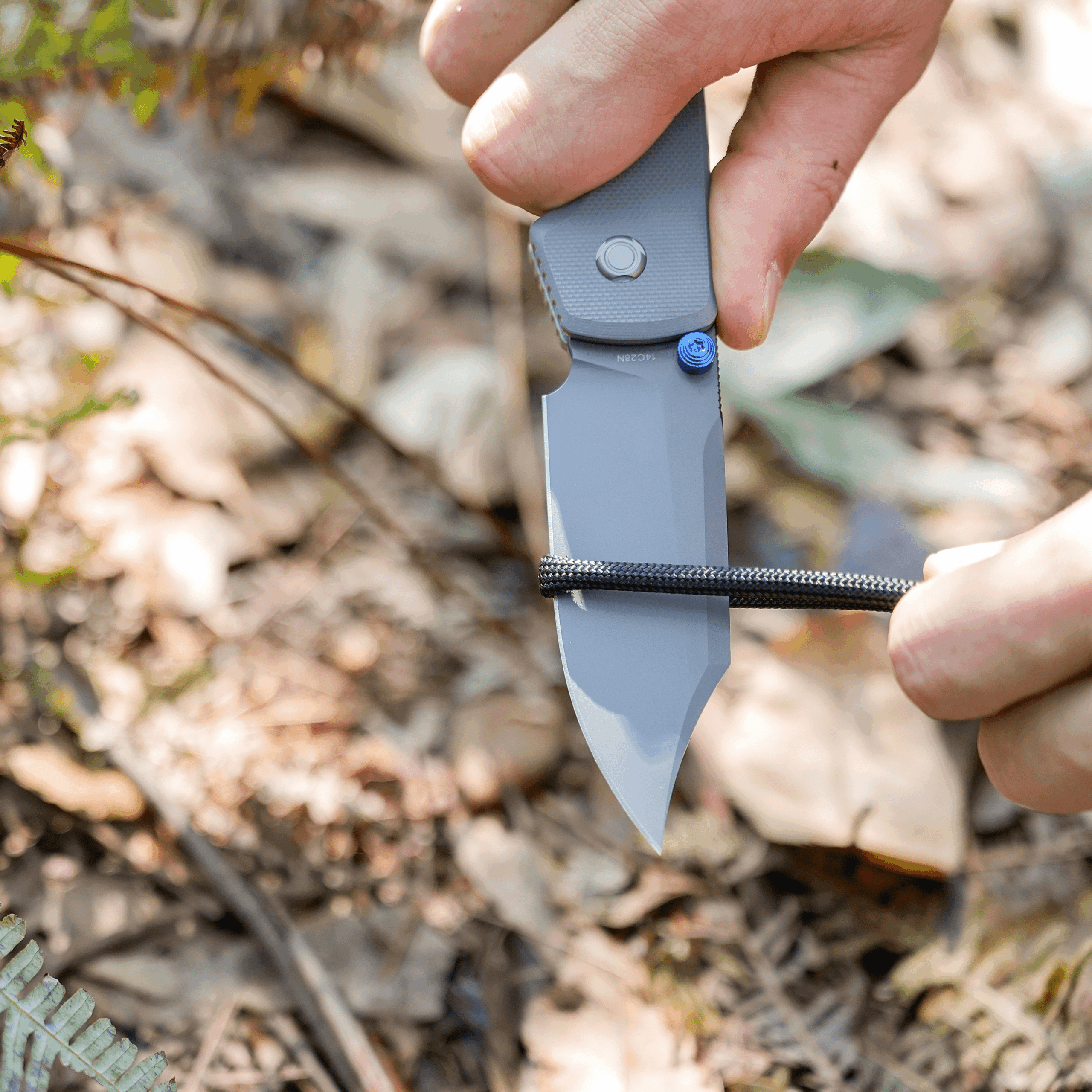 Close-up of a hand gripping a gray folding knife outdoors, blade visible against a background of leaves and dirt.