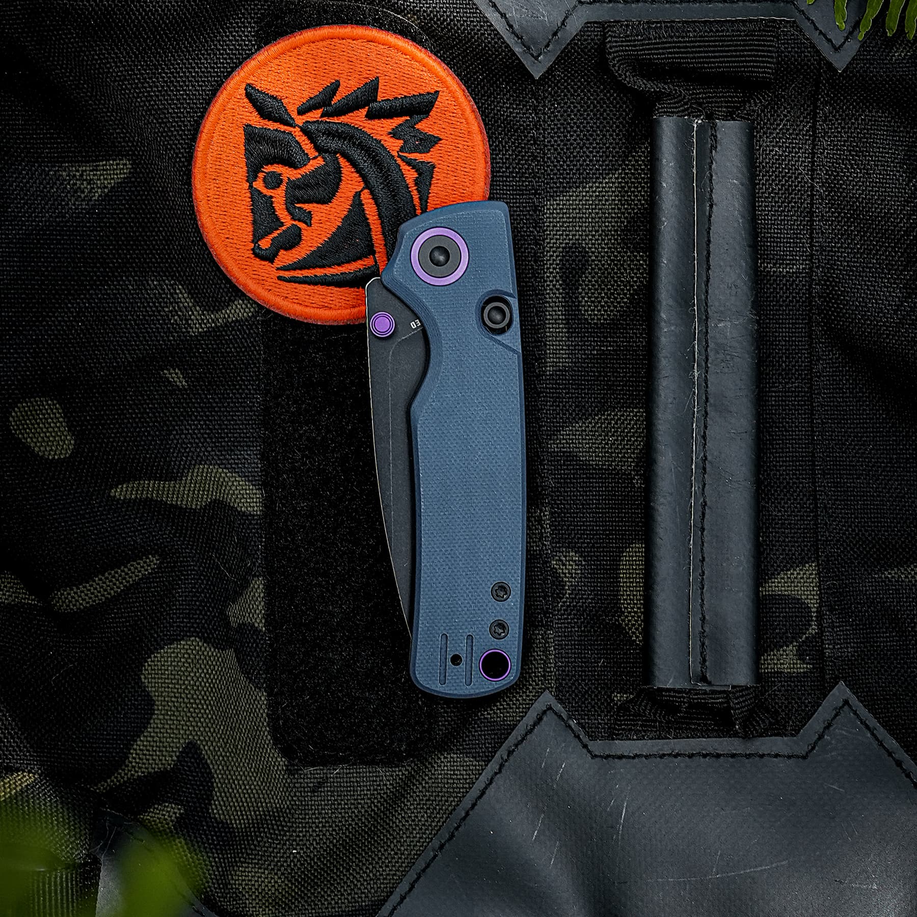 Close-up of a tactical backpack with an orange circular patch featuring a black stylized dragon logo and a blue folding knife with purple accents attached to the front.