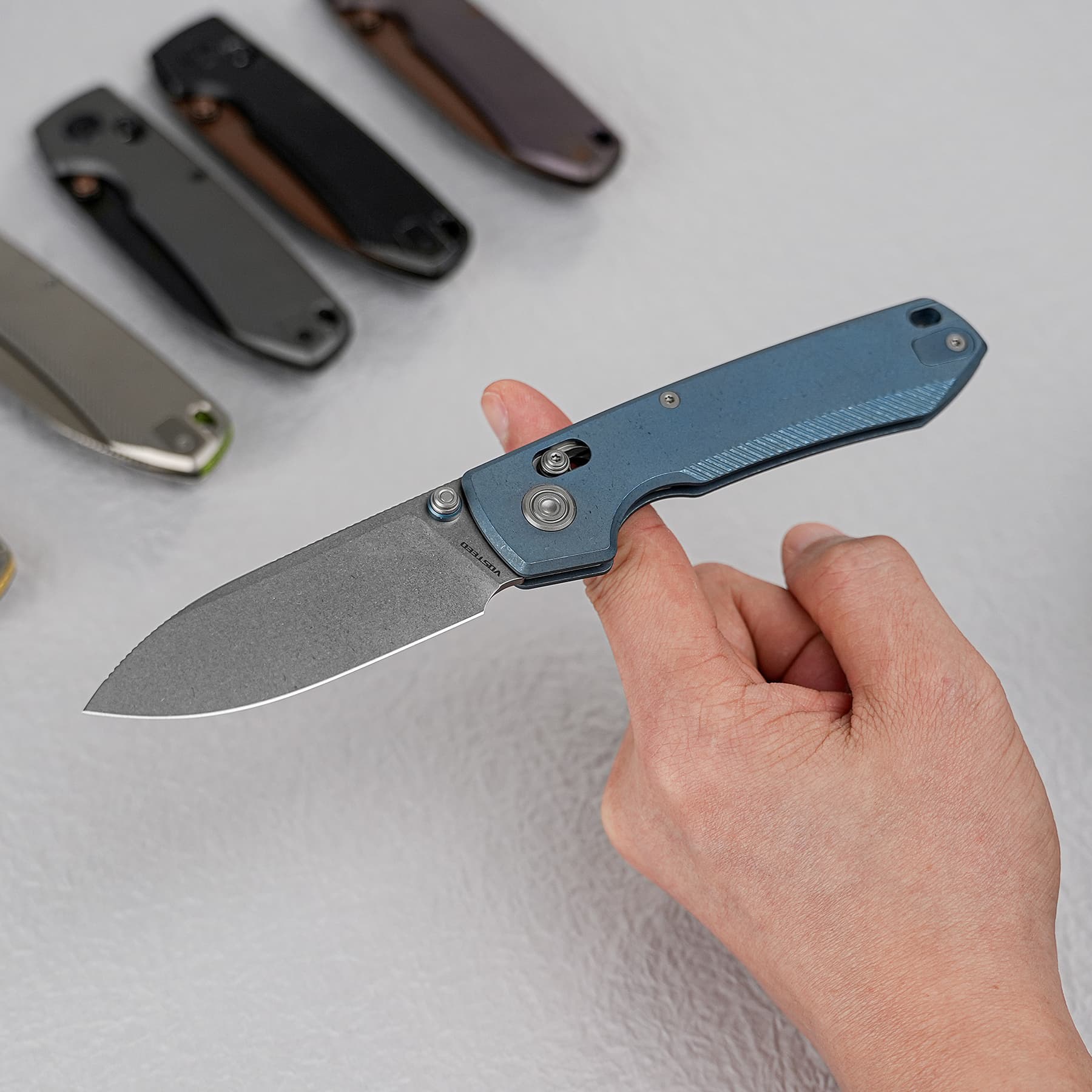 Blue folding knife held in hand; four other knives blurred in the background on a gray surface, foreground shows blade edge and screws.