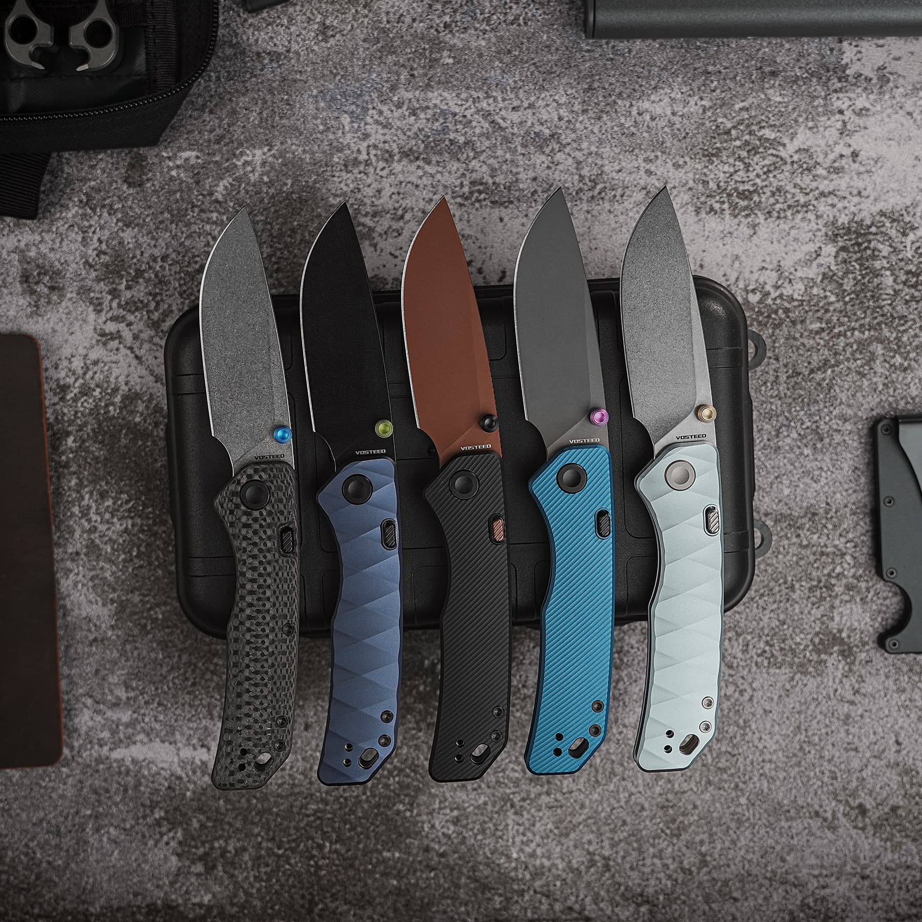 Five folding knives with colored handles (blue, black, brown, gray, light blue) in a black case on a textured surface.