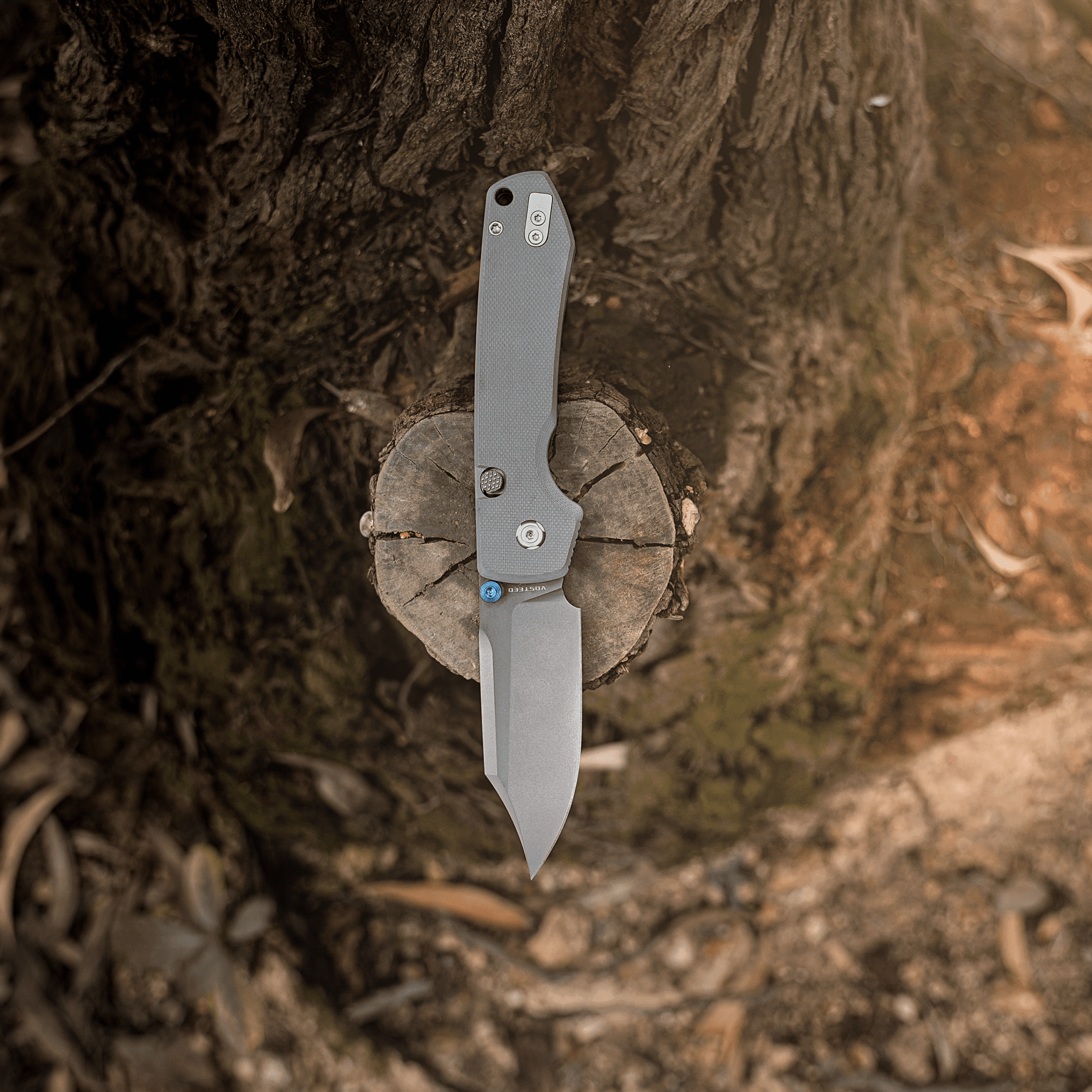 Gray textured folding knife embedded in a tree trunk, blade partly visible and pointing downward.