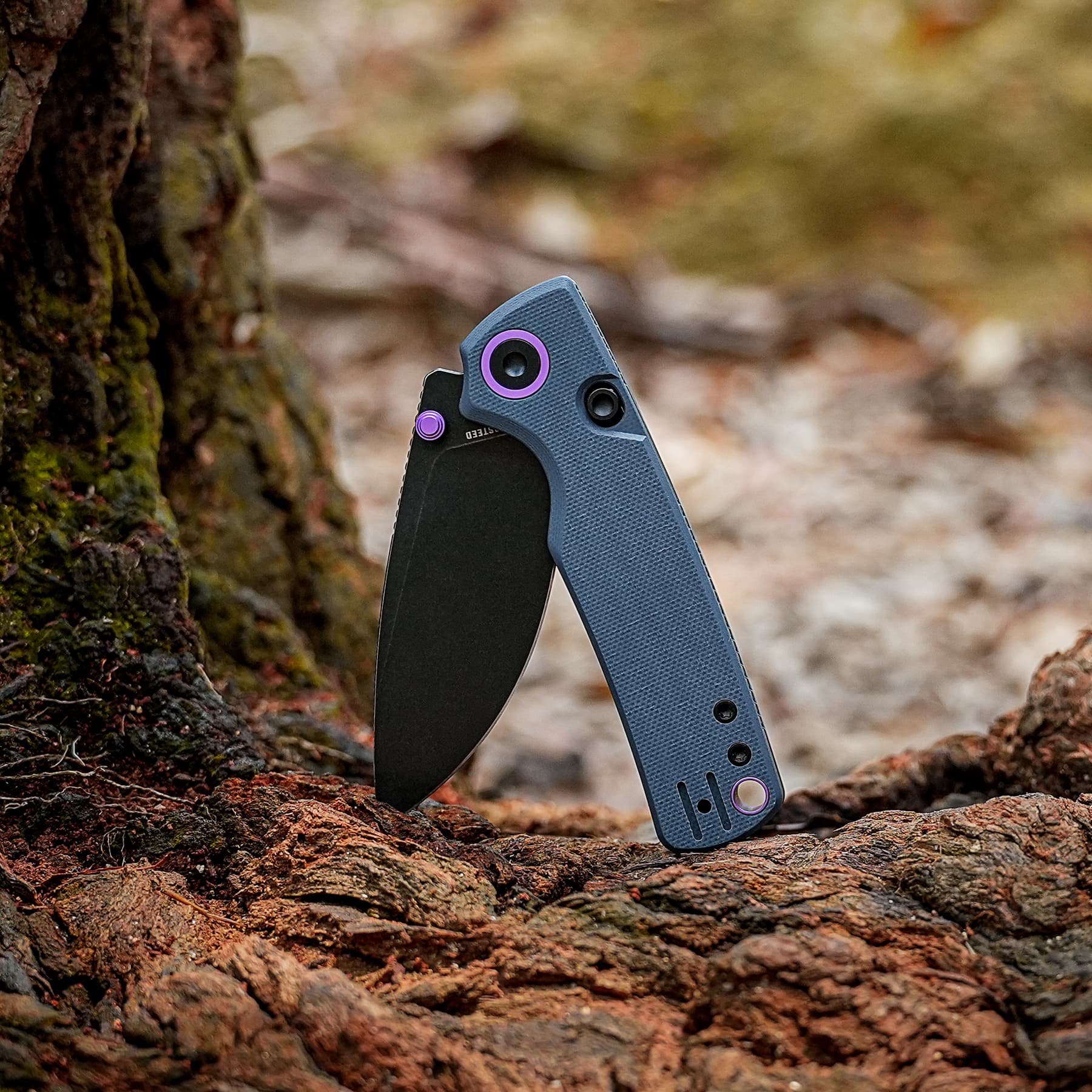 Blue folding knife with purple accents wedged between rocks in an outdoor setting.