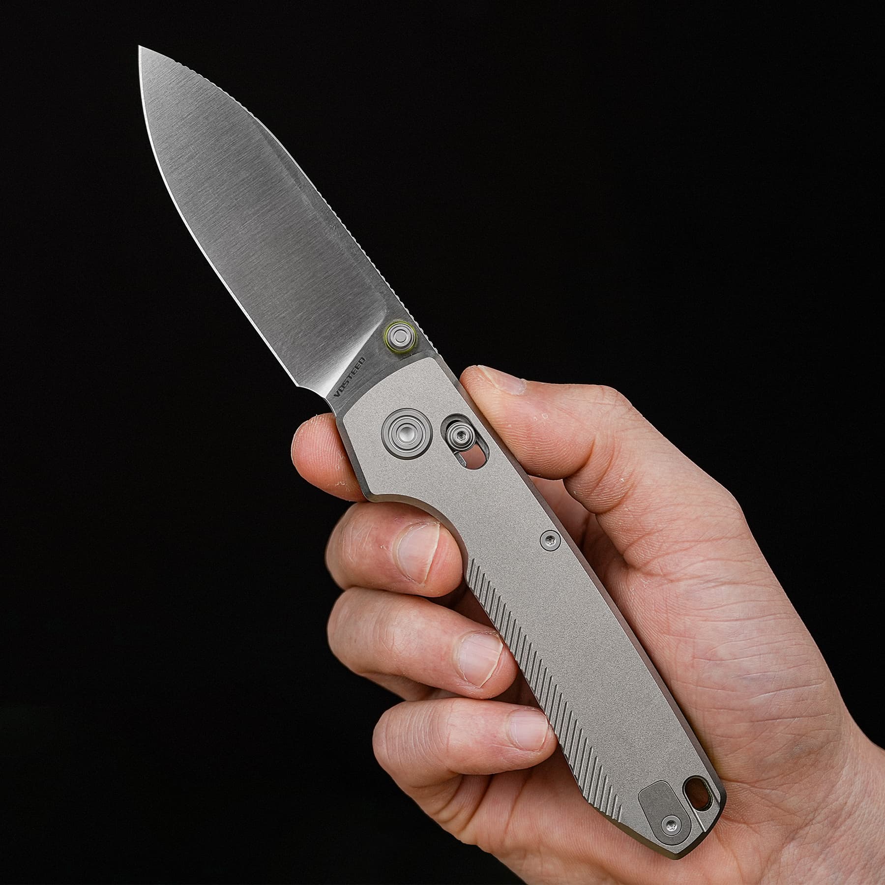 Close-up of a hand gripping a gray folding knife with a stainless blade against a black background.