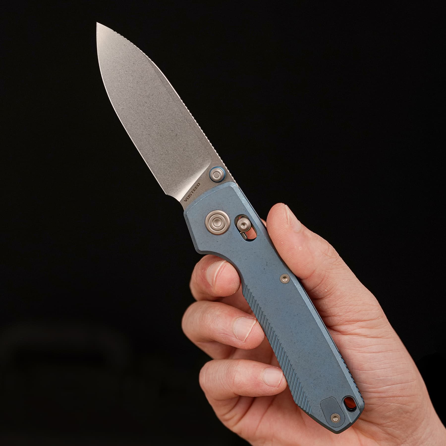 Close-up of a folding knife blade held in a hand against a black background, blade pointed upward to left.