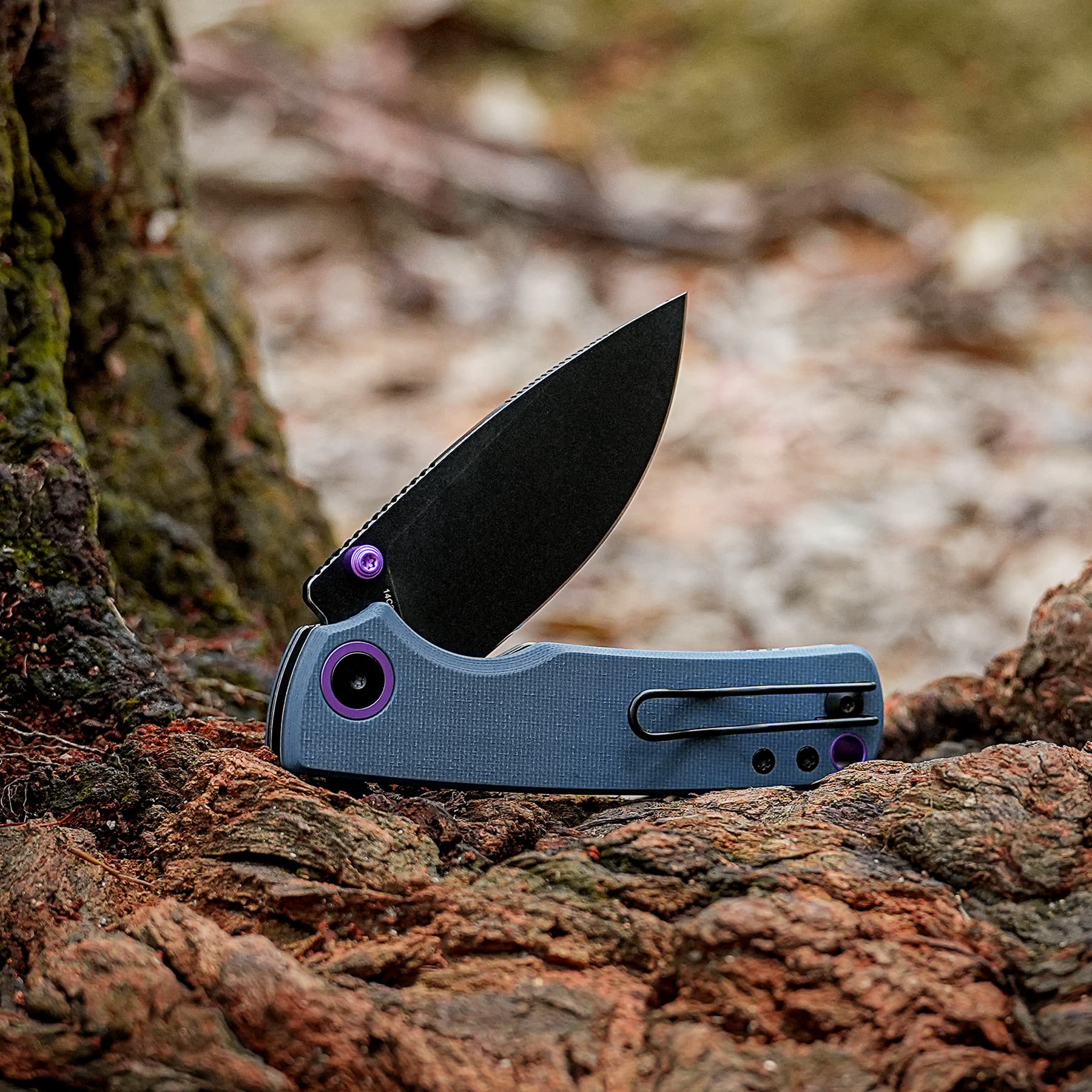 Black folding knife partially embedded in rough reddish-brown rocks outdoors, purple hardware visible on the handle edge
