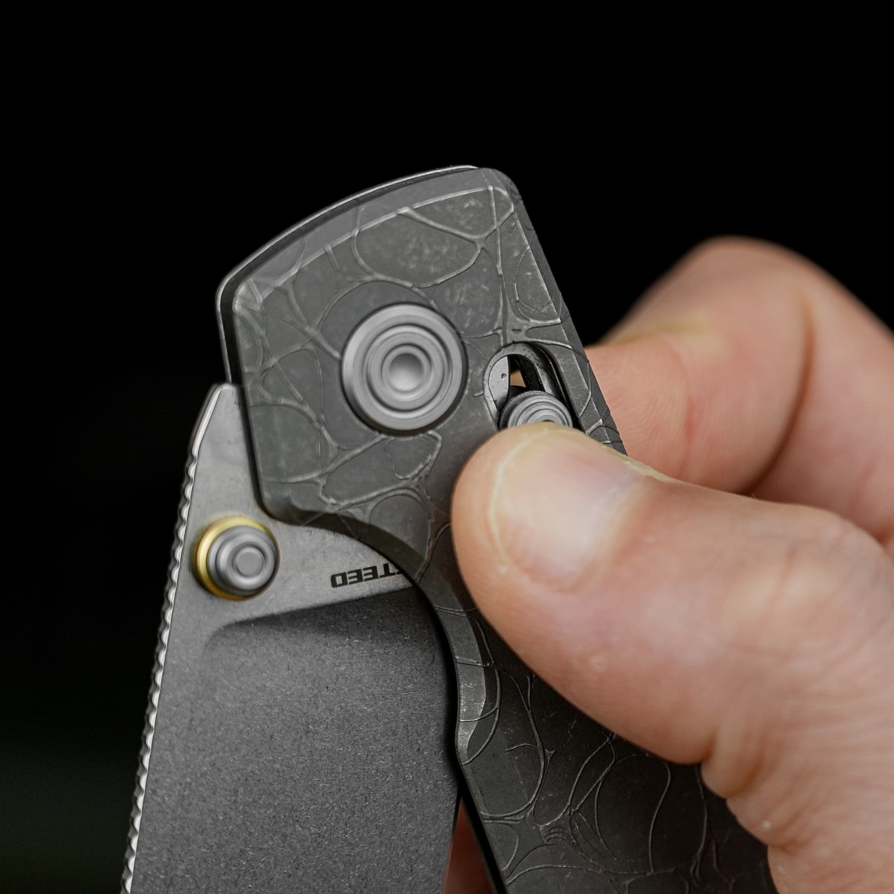 Close-up of a hand holding a gray folding knife blade with a textured handle and circular thumb studs against a black background.