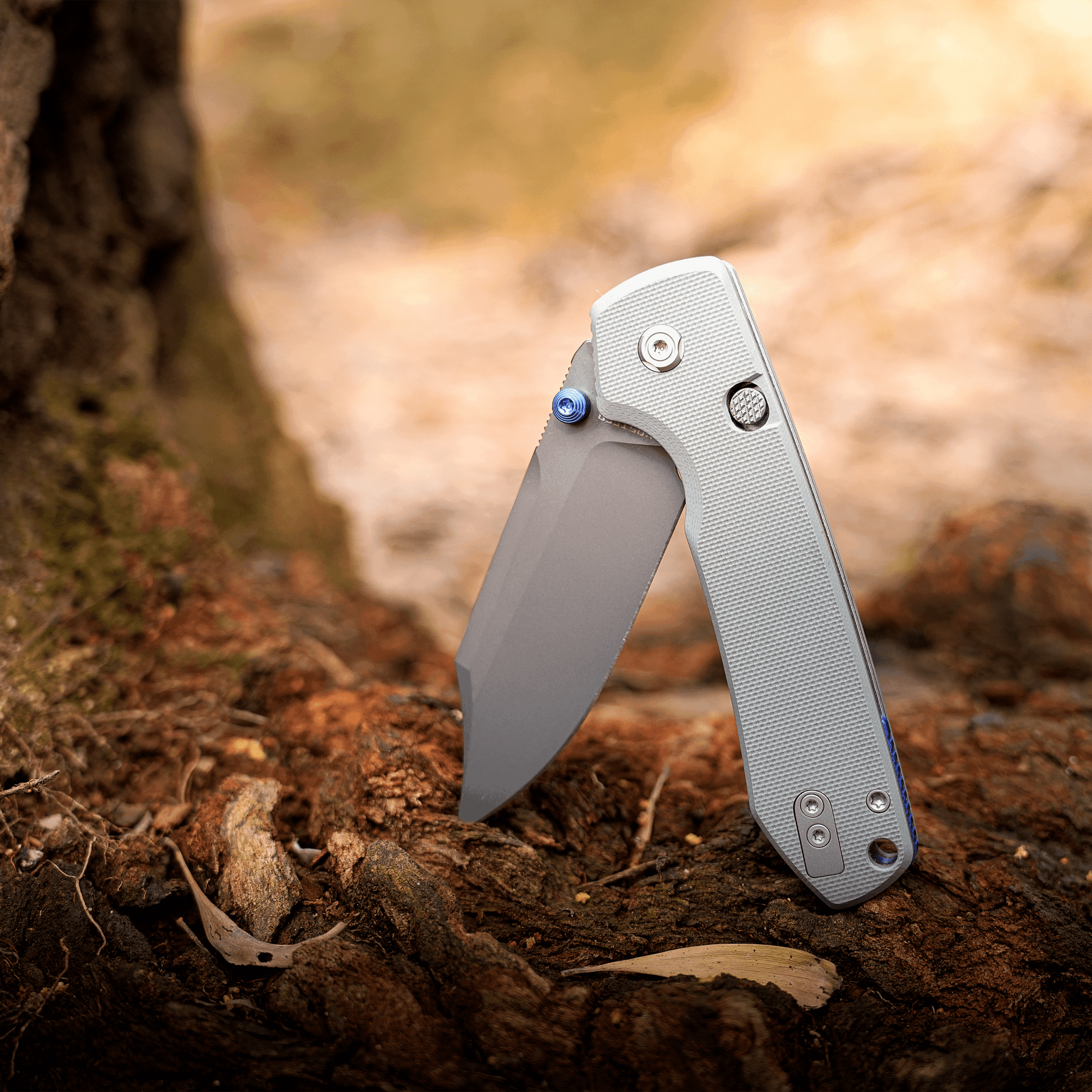 Close-up of a gray folding knife blade partially opened, stuck in loose dirt outdoors.