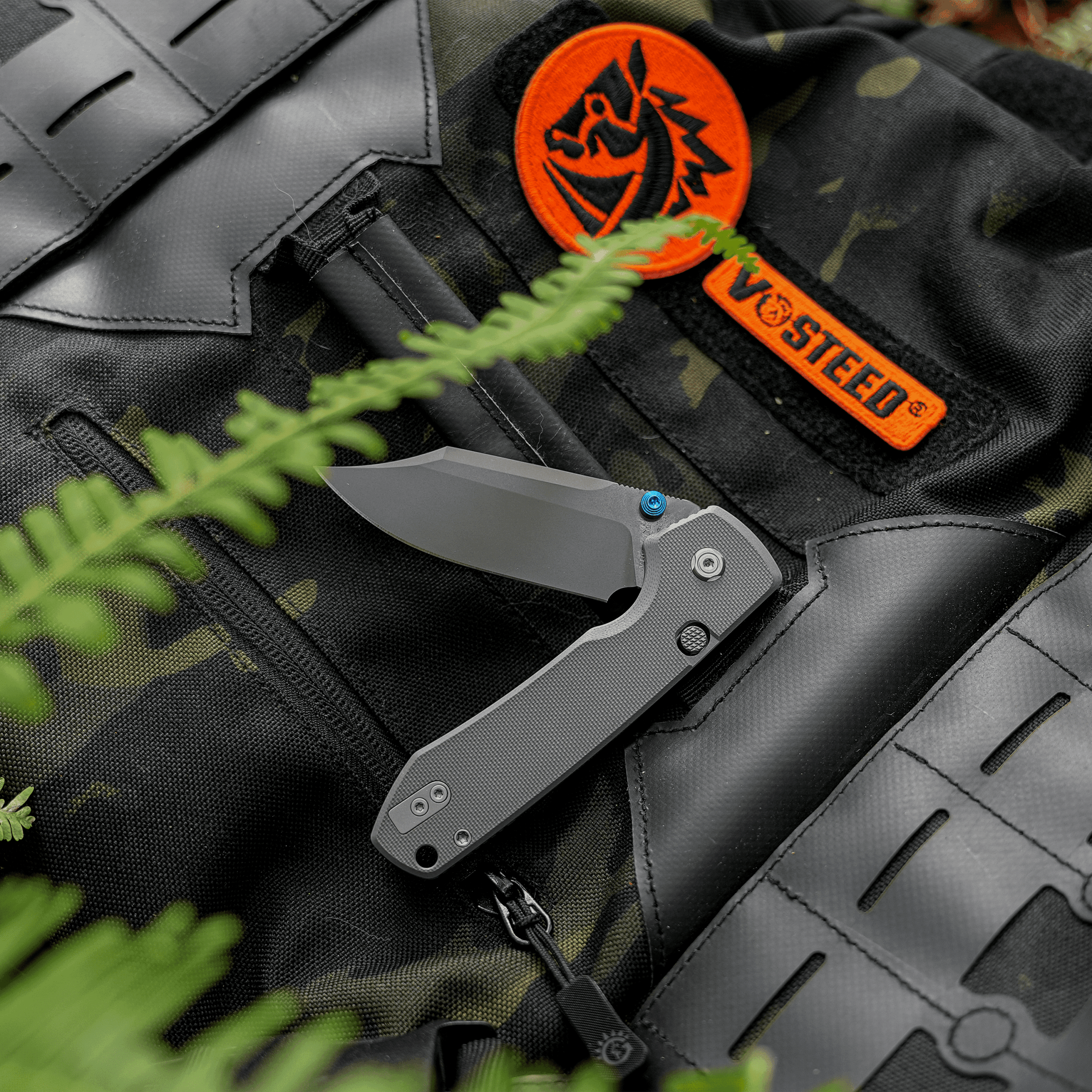 Close-up of a gray folding knife resting on a camouflage tactical vest, with an orange circular patch featuring a black emblem and an orange strap partially visible.