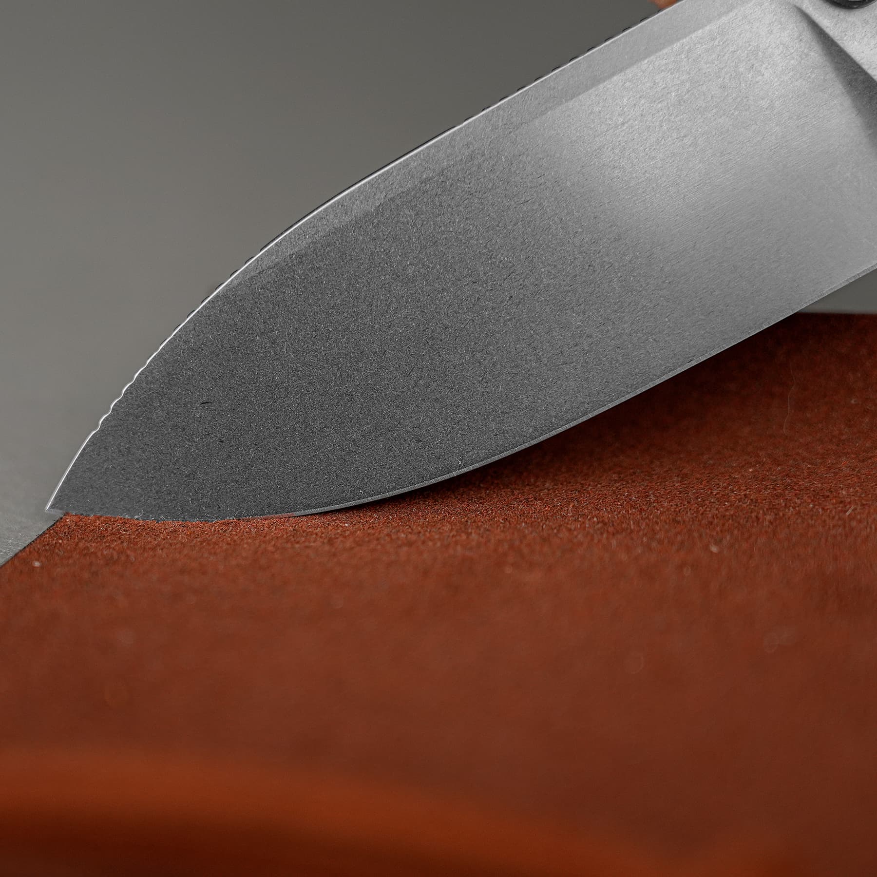 Close-up of a gray serrated knife blade resting on a reddish-brown surface, showing the curved edge and texture of the metal.