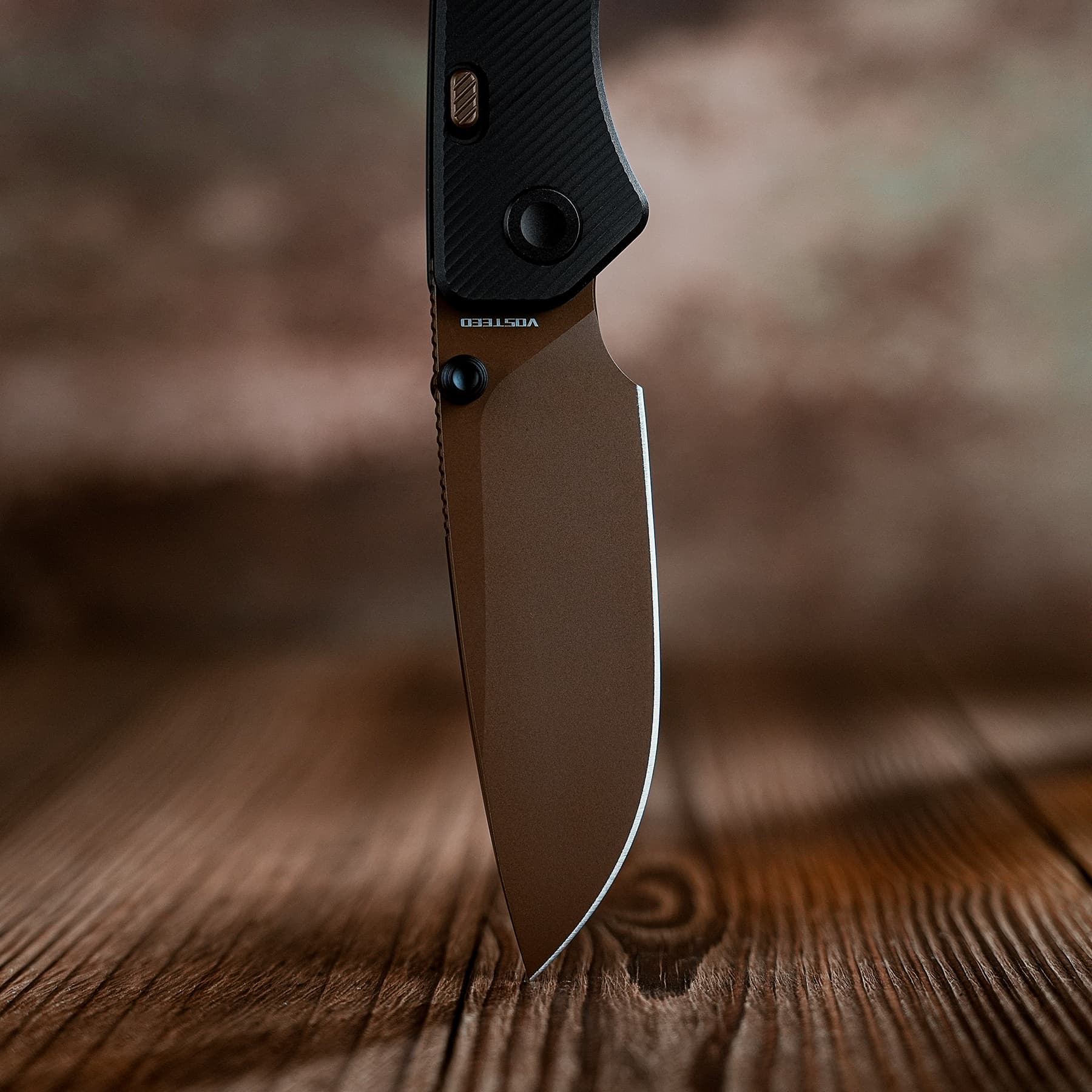 Close-up of a folding knife blade meeting its textured black handle with a wooden surface beneath, showing a browns/blacks color and a thumb stud.