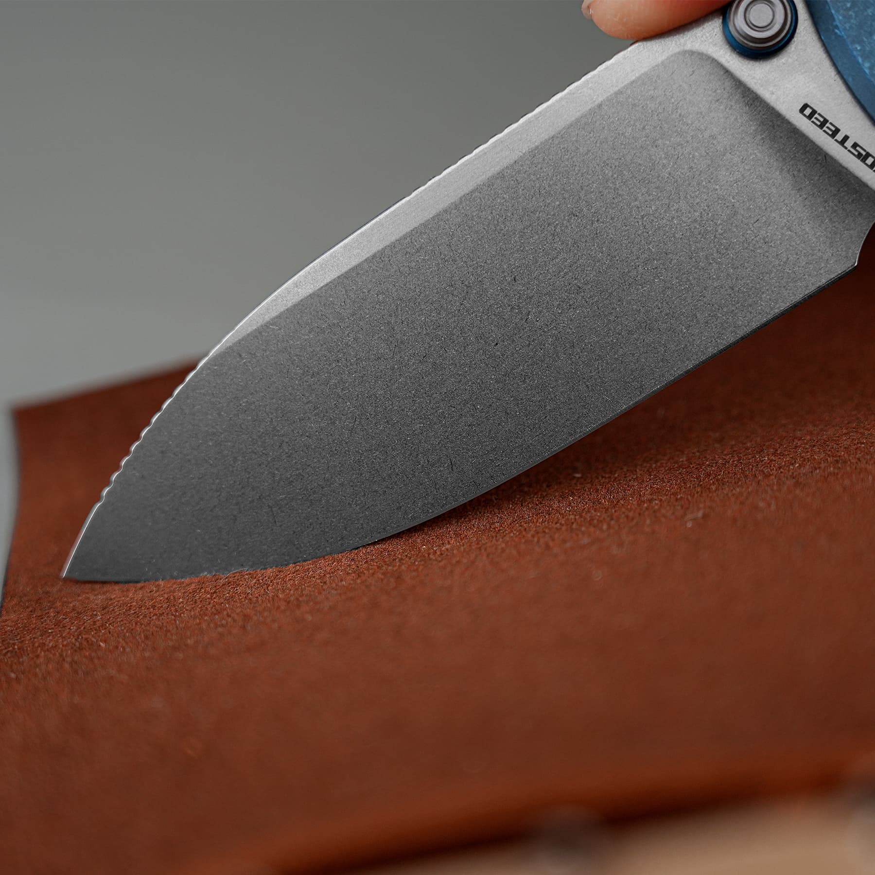 Close-up of a knife blade angled across a brown textured surface, showing the sharpened edge and part of the blue-handle near the pivot.