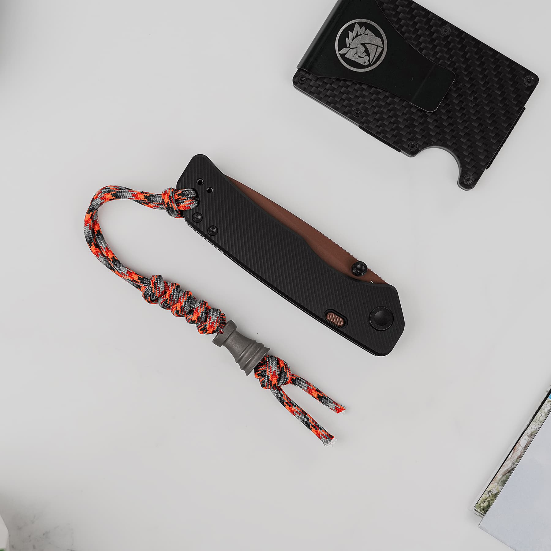 Close-up of a black folding knife with a textured handle, attached to a red‑gray paracord lanyard, on a light gray surface; a carbon‑fiber sheath is in the background.