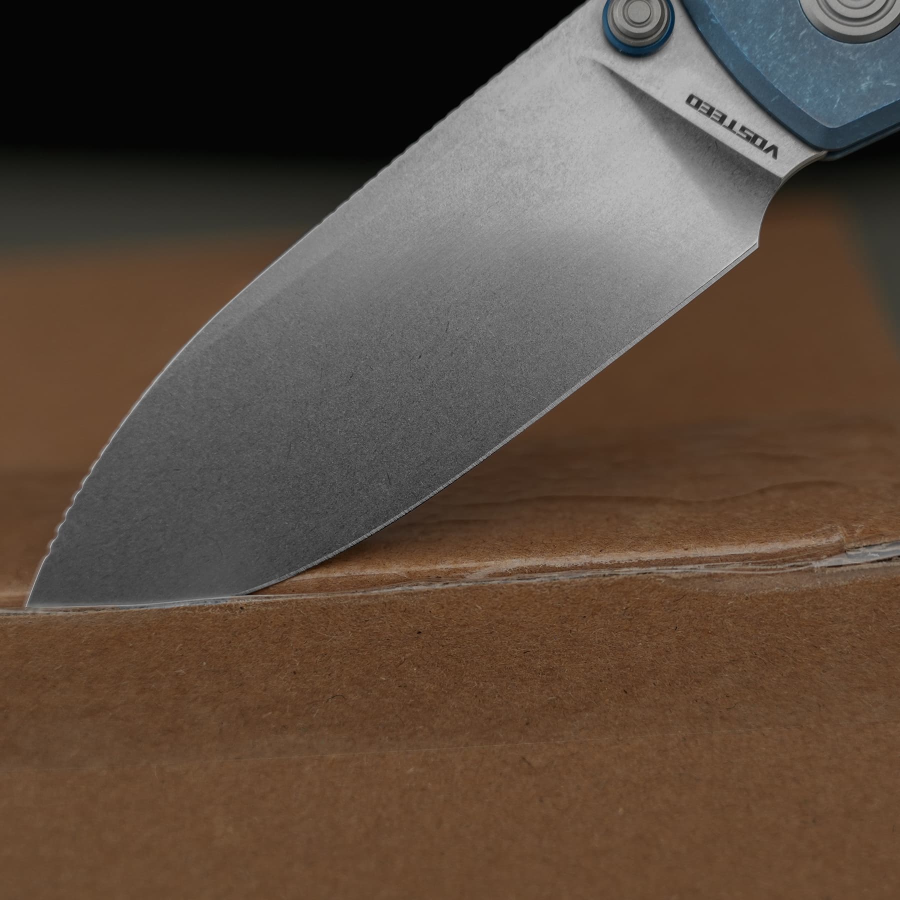 Close-up of a folding knife blade angled over a wooden table, showing a worn edge and part of the blue handle.